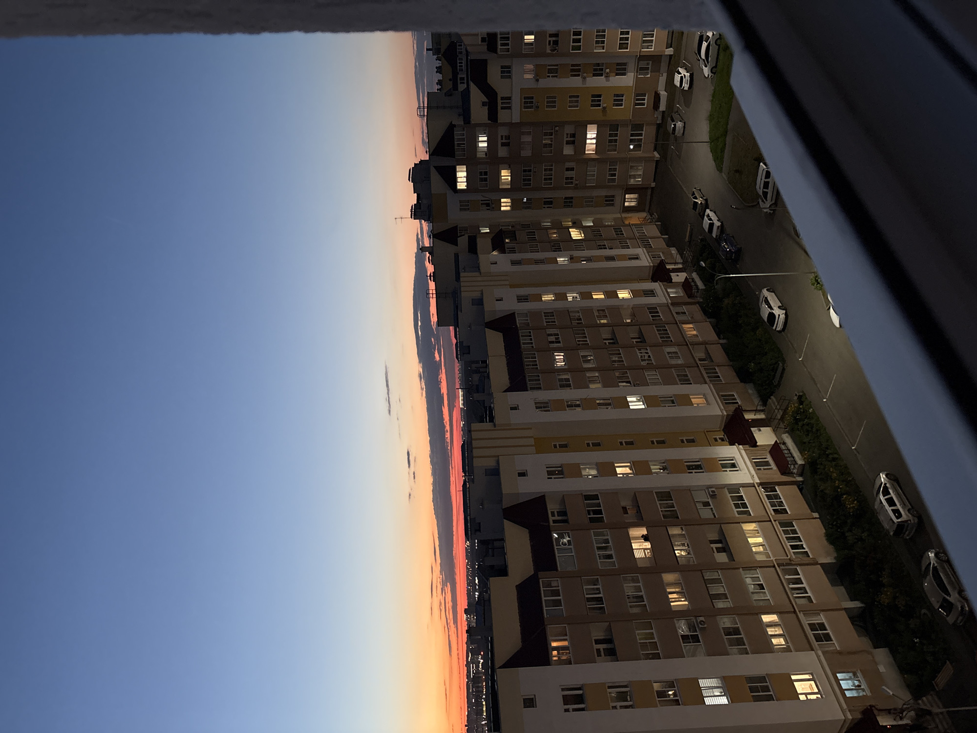 This is a high-angle, outdoor, vertical shot of an apartment complex at dusk in an urban environment. The sky is a gradient of deep blue transitioning to a fiery orange and red band of sunset on the horizon, with scattered dark clouds. The apartment buildings are tall, multi-story structures with numerous windows, many of which are illuminated from within, suggesting it is evening. The architecture appears to be modern, with a mix of light-colored facades and yellow accent details. Below the buildings, a paved courtyard area is visible with several parked cars, including a silver sedan, a silver SUV, and several white cars. Green grass and some vegetation are present in small patches around the buildings and courtyard. The image is framed by a dark, textured surface in the foreground, possibly a windowsill or balcony railing.