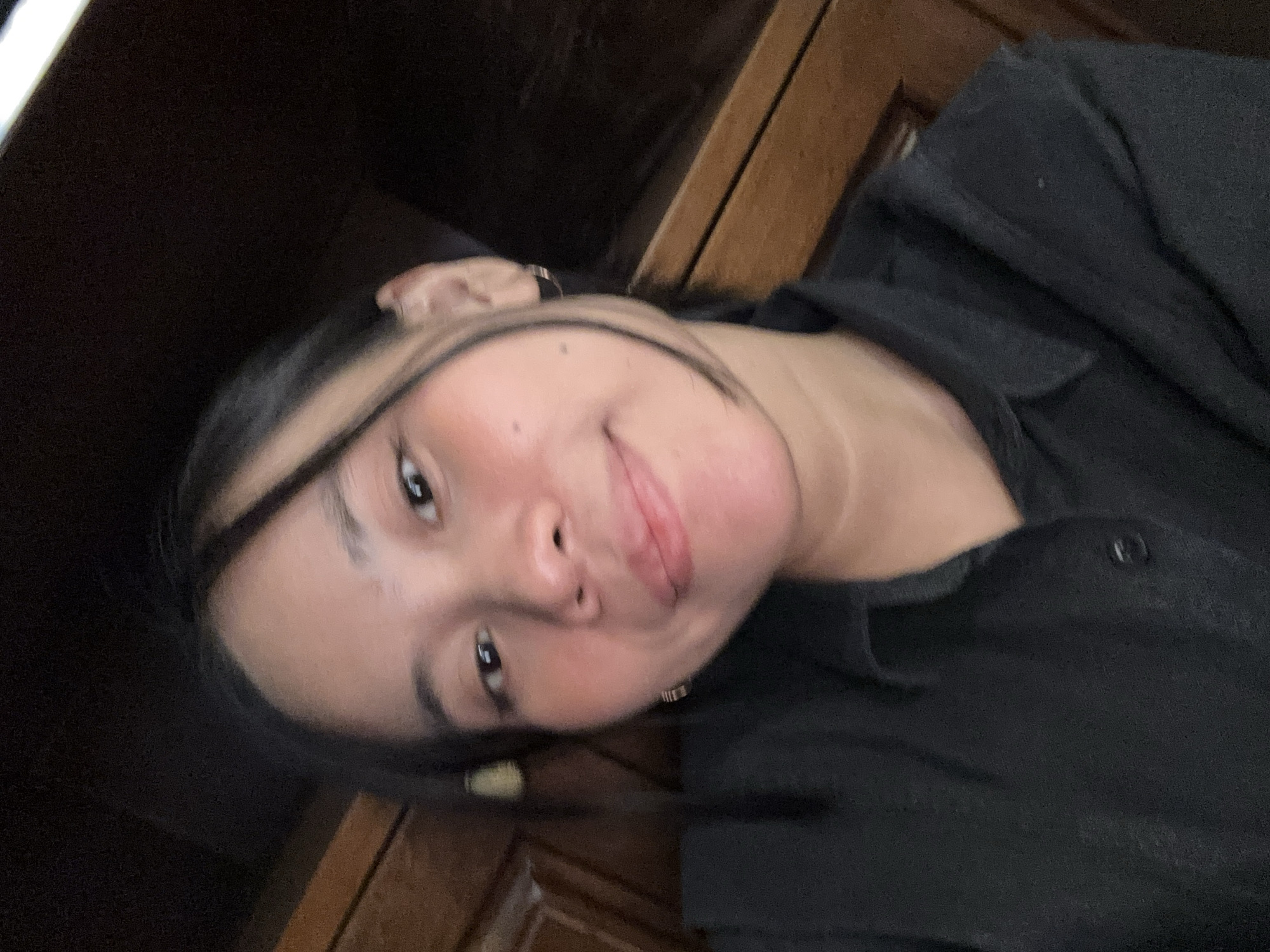 A young woman with dark hair and fair skin is taking a selfie in a room with wooden paneling. She is wearing a black collared shirt. Her hair is pulled back, with some strands framing her face. She has a light smile and her eyes are looking towards the camera. The background appears to be a wall with horizontal wooden planks. The image is lit with soft light, possibly from a window or overhead lighting. There is no indication of the specific location within South Jakarta, Indonesia, beyond the presence of a residential or indoor setting.