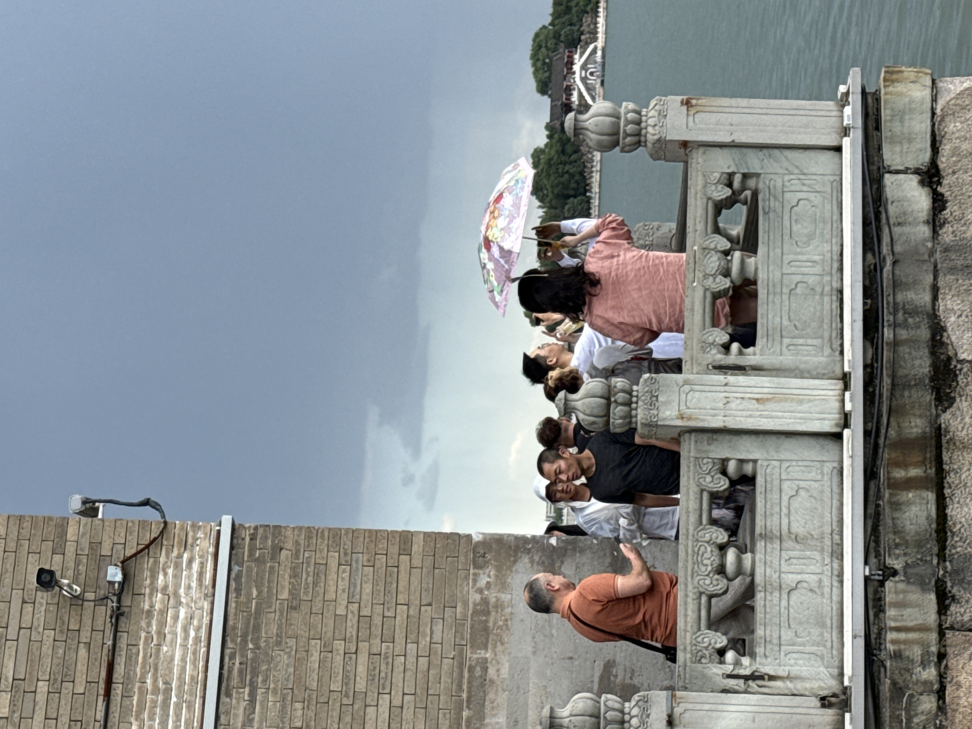 The image depicts a crowded outdoor scene on a stone balustrade overlooking a large lake in the Haidian District of Beijing, China, specifically within the Summer Palace (Yiheyuan) complex, likely facing Kunming Lake towards Nanhu Island.
In the foreground to the left, a weathered light-brown brick wall is visible. Mounted on this wall are two security cameras: a white dome camera facing downwards and a silver bullet-style camera extending horizontally, both connected by visible conduits and wiring, some of which show signs of rust. Below the brick, the wall transitions to a lighter, stained stone or concrete base.
A grey stone balustrade, intricately carved with ornate pillars featuring vase-like tops and decorative panels with cloud or scroll motifs, spans across the lower half of the frame. Behind this balustrade, a group of approximately eight people, primarily adults, are gathered.
From left to right along the balustrade:
* A Caucasian male in an orange short-sleeved shirt with a black shoulder bag is speaking to another man. His left hand is slightly raised in a gesture.
* Next to him, an Asian male in a dark grey short-sleeved shirt stands with his back partially turned, looking towards the lake.
* Partially obscured behind them is an individual in a white hooded garment.
* Further to the right, an Asian female in a light pink short-sleeved top holds a colorful umbrella with a floral pattern over her head, using both hands to photograph or video the distant scenery with a smartphone.
* Other individuals are visible to her left and right, also observing the view, with at least one more person holding a smartphone.
The background features a vast expanse of calm, greenish-blue water, which is Kunming Lake. On the far side of the lake, a traditional Chinese architectural complex stands on a low, tree-covered landmass, consistent with Nanhu Island. The prominent building has dark roofs, white walls, and a distinctive arched entrance or bridge leading up to it, surrounded by lush green trees. The sky above is overcast with dark grey clouds, indicating cloudy weather.