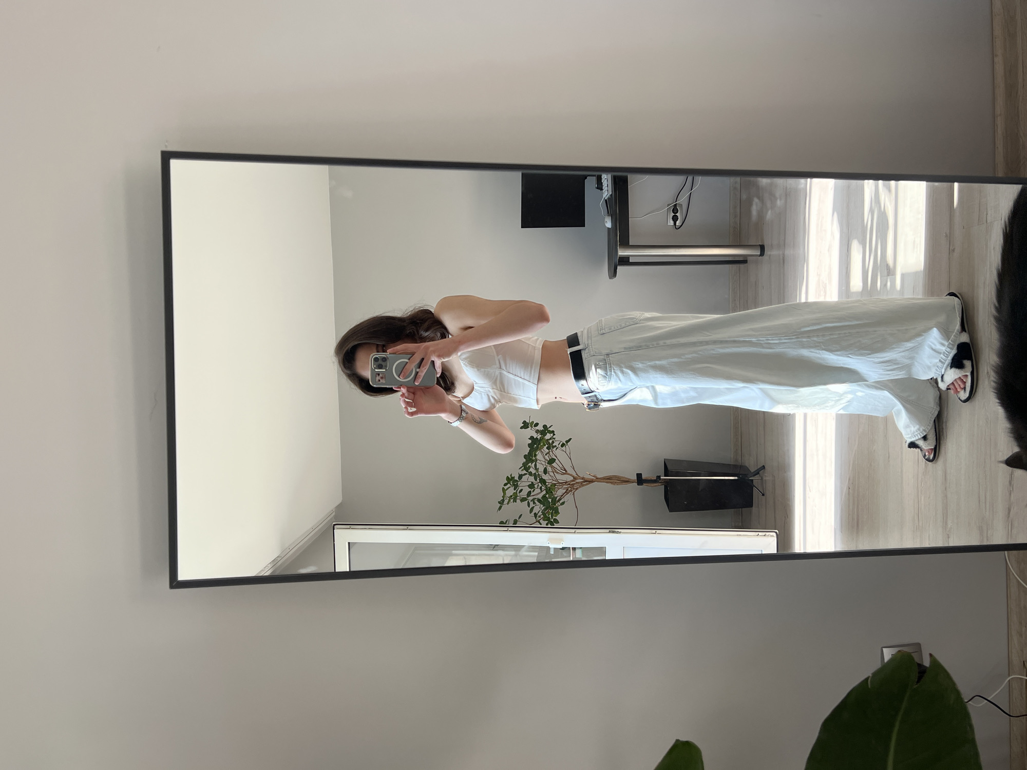 A young woman stands in front of a full-length mirror in what appears to be a residential interior. She is taking a selfie with her phone, holding it up to capture her reflection. She is wearing a white corset-style top, light blue wide-leg jeans, a black belt, and fluffy black and white slide sandals. Her dark brown hair is shoulder-length and styled in loose waves.
The mirror is tall and rectangular with a black frame, placed against a light gray wall. In the reflection, a doorway with a glass panel is visible, leading to an unseen space. To the right of the doorway, a tall potted plant with green leaves stands in a black planter. Further to the right, a black desk with a computer monitor and keyboard is visible. Sunlight streams in from an unseen window, casting shadows on the light wooden floor. The room is sparsely furnished, suggesting a minimalist aesthetic. There is no indication of the location being in Ternopil, Ukraine, nor any other specific city.