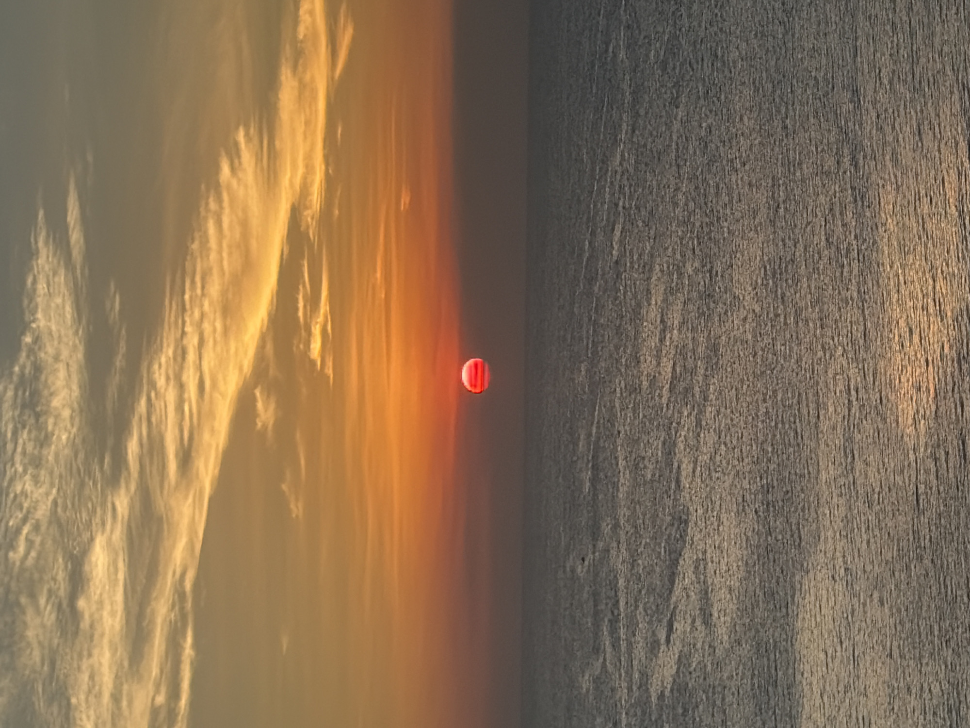 A vivid sunset over a calm ocean is depicted. The sun, a deep crimson sphere, is positioned at the horizon line, casting a warm glow across the sky. Wispy clouds, tinged with orange and yellow, stretch across the upper half of the frame, creating a dramatic backdrop. The sea below reflects the sunset's colors, with subtle ripples disturbing the otherwise placid surface. A faint silhouette of what appears to be a bird can be seen in the distance, adding a touch of life to the vast expanse. The overall impression is one of serene natural beauty.