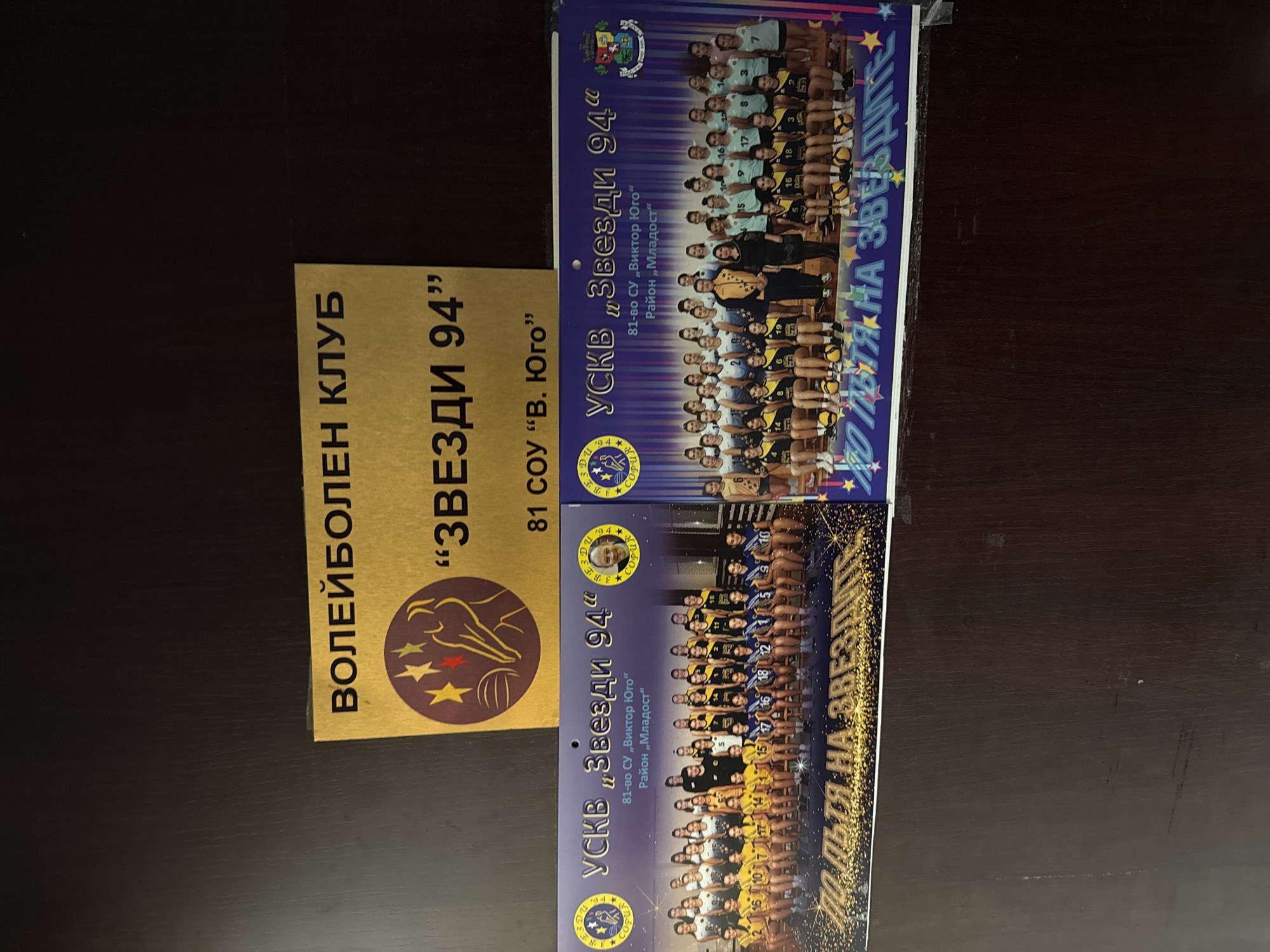 The image displays three printed items on a dark, textured surface, likely a desk or shelf, under a darker background. A rectangular golden-yellow plaque is positioned centrally at the top, featuring the text "ВОЛЕЙБОЛЕН КЛУБ" (Volleyball Club) and "„ЗВЕЗДИ 94”" ("ZVEZDI 94" / Stars 94) in black Cyrillic letters. Below this, smaller text reads "81 СОУ “В. Юго”" (81 Secondary School "V. Yugo" / Viktor Hugo) and "Район „Младост”" (Mladost District). A maroon circular logo with a stylized hand reaching for three stars (yellow, red, green) is present on the plaque.
Below the plaque, two rectangular, glossy team photographs are laid side-by-side. Both photographs feature the title "УСКВ „Звезди 94“" (USKV "Zvezdi 94"), along with "81-во СУ „Виктор Юго“" (81st Secondary School "Victor Hugo") and "Район „Младост“" (Mladost District), indicating the location as the 81st Secondary School "Victor Hugo" in the Mladost district of Sofia, Bulgaria.
The left photograph depicts approximately 26 young females, presumably volleyball players, posed in rows on a court-like surface. They wear various team uniforms in yellow, black, white, and blue. Two adult figures, one male in a yellow and black tracksuit and one female in a black dress, stand centrally among the players. Several volleyballs are placed on the ground in front of the sitting players. The bottom edge of the photo features glittery text "ПО ПЪТЯ НА ЗВЕЗДИТЕ" (On the Path of the Stars). Circular club logos are visible in the top corners, with one containing a small portrait of a male.
The right photograph similarly displays approximately 28 young females, also volleyball players, in rows on a court. They wear light blue and black team uniforms. The same male coach in the yellow and black tracksuit stands centrally, accompanied by another adult female in a black dress. Volleyballs are visible on the ground. This photo also includes the "ПО ПЪТЯ НА ЗВЕЗДИТЕ" text along its bottom edge. Circular club logos appear in the top corners, and a municipal coat of arms is visible in the top right corner.