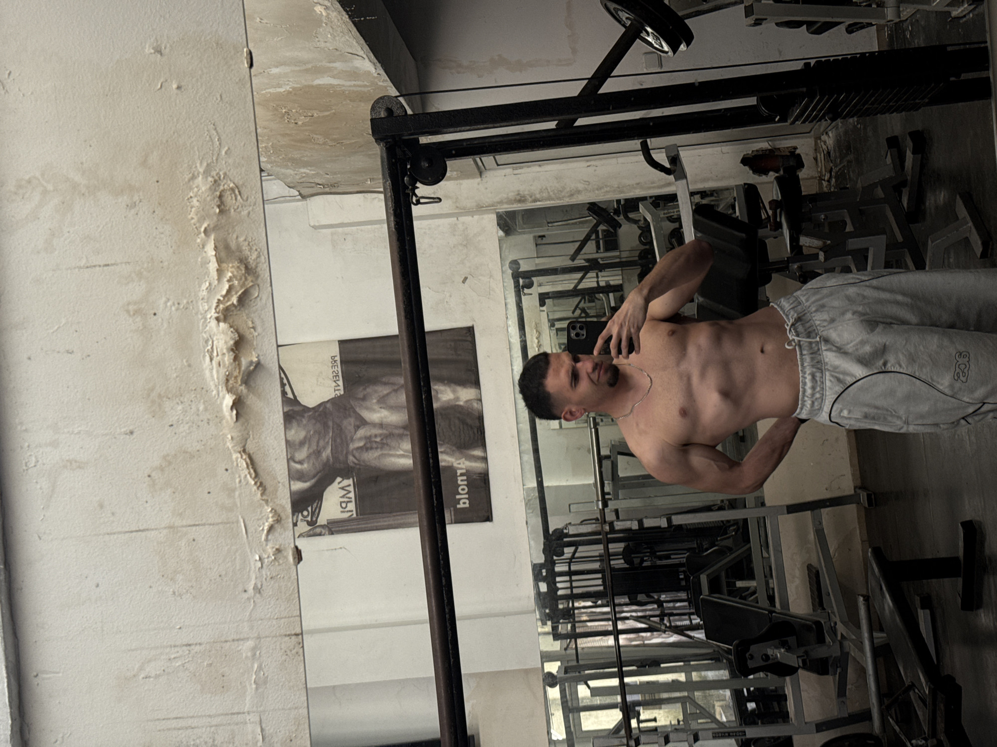 A sideways-oriented indoor photo captures a shirtless young man posing in a gym in Shumen, Bulgaria. The man, with short dark hair and a beard, is positioned on the right side of the frame, looking towards the top-left while holding a black smartphone to his ear. He wears light grey sweatpants featuring a small, indistinct logo on the lower-right leg, and a thin chain necklace. His physique appears well-defined.
A large, somewhat aged mirror occupies the lower portion of the image, reflecting various pieces of dark, industrial-style gym equipment like weight machines, racks, and benches, as well as the man’s reflection.
The left and upper parts of the image reveal a light-colored wall, which is notably damaged. Extensive peeling paint, water stains, and discoloration are visible, especially in the upper-left and top sections. On this wall, a large black and white poster depicts a muscular male torso. Visible text on the poster, which appears reversed due to its orientation in the image, includes "IMY", "blonm", and Cyrillic characters "ББЕ2ЕН". The ceiling, visible in the background, also shows signs of wear.
The overall atmosphere suggests an older or less recently renovated fitness facility. The time of day is not clearly indicated but the lighting implies an indoor, possibly artificial, setting.