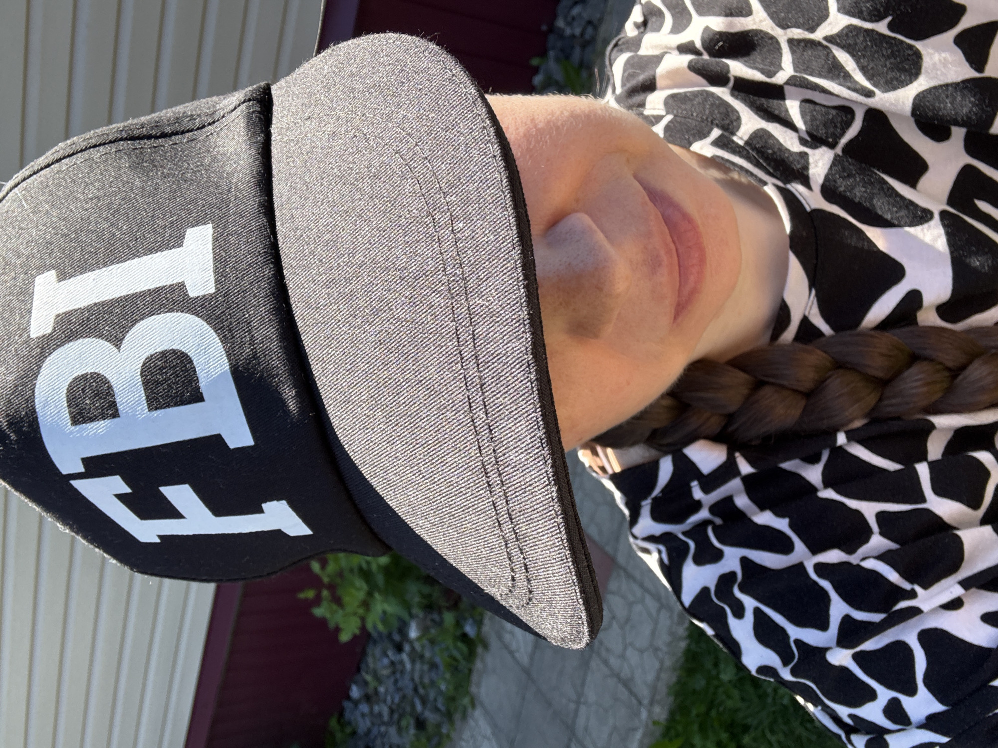 A young Caucasian woman is captured from the chest up, wearing a black baseball cap with white "FBI" lettering. The cap's brim casts a shadow over her eyes. She has a single braid of brown hair extending over her shoulder and is wearing a black and white patterned shirt. The background shows the exterior of a building with horizontal siding and a dark red lower portion, adjacent to a gravelly area with green foliage and a paved walkway. The scene is lit by direct sunlight.