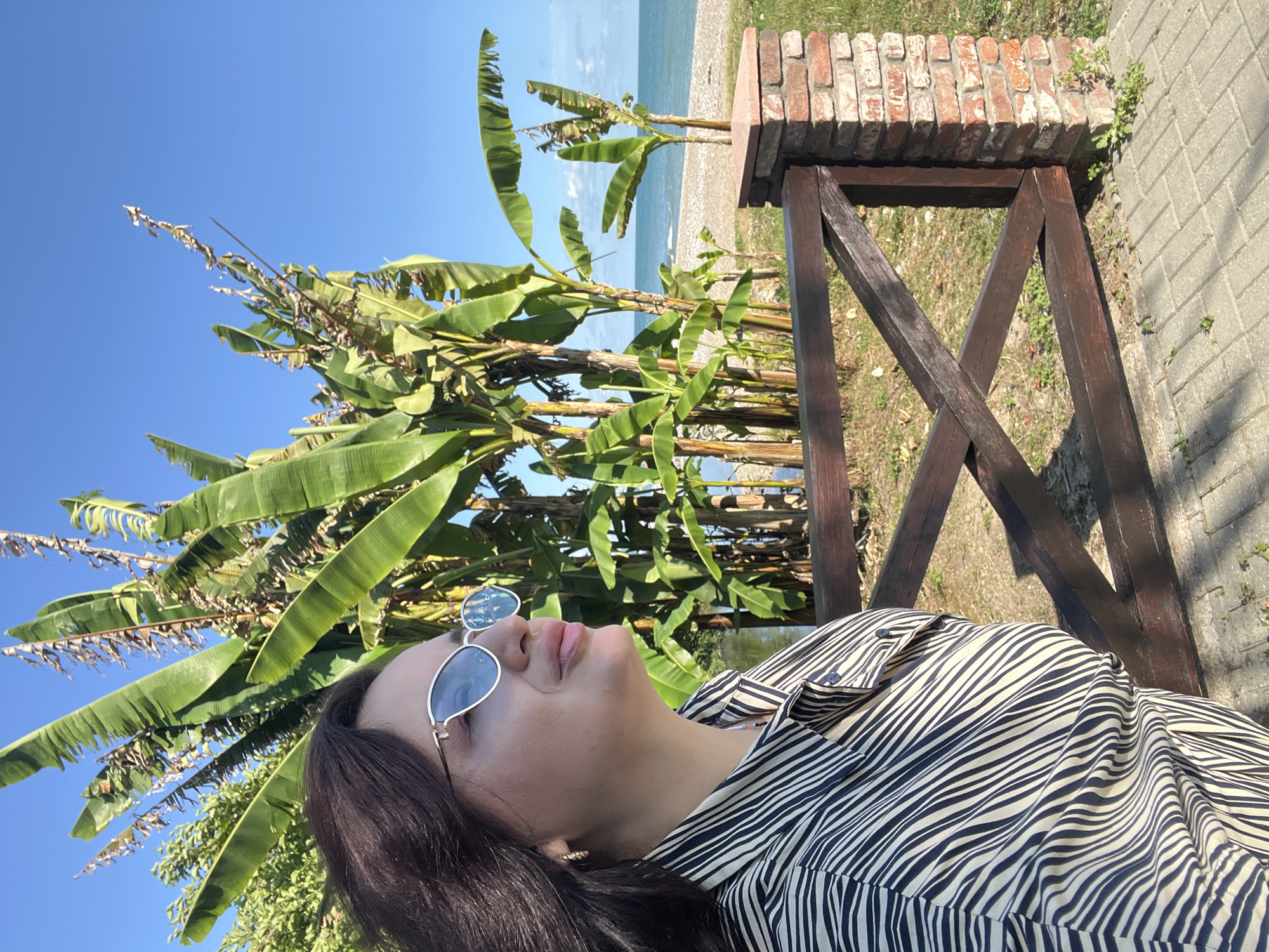 A medium shot captures a woman with dark hair, wearing sunglasses and a black and white striped shirt, standing outdoors. She is positioned in the foreground, her body angled to the right, and her head is turned slightly to the right, looking upwards and to the right. Behind her, a vibrant blue sky is visible, with lush green banana trees creating a tropical backdrop. A wooden fence with a brick pillar is in front of her, and beyond that, a pebbled beach meets the calm blue sea. The lighting suggests it is daytime.