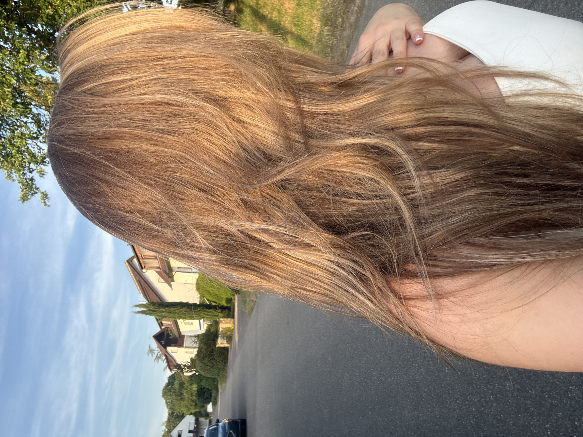 A person's long, blonde hair is in the foreground, obscuring their face. The person is wearing a white top. Behind them, a paved road curves to the left, lined with houses and green trees. A dark-colored car is parked on the side of the road on the left. The sky is blue with wispy clouds, and the sun is setting, casting a warm glow on the scene. The overall impression is of a quiet suburban street bathed in the golden light of dusk.