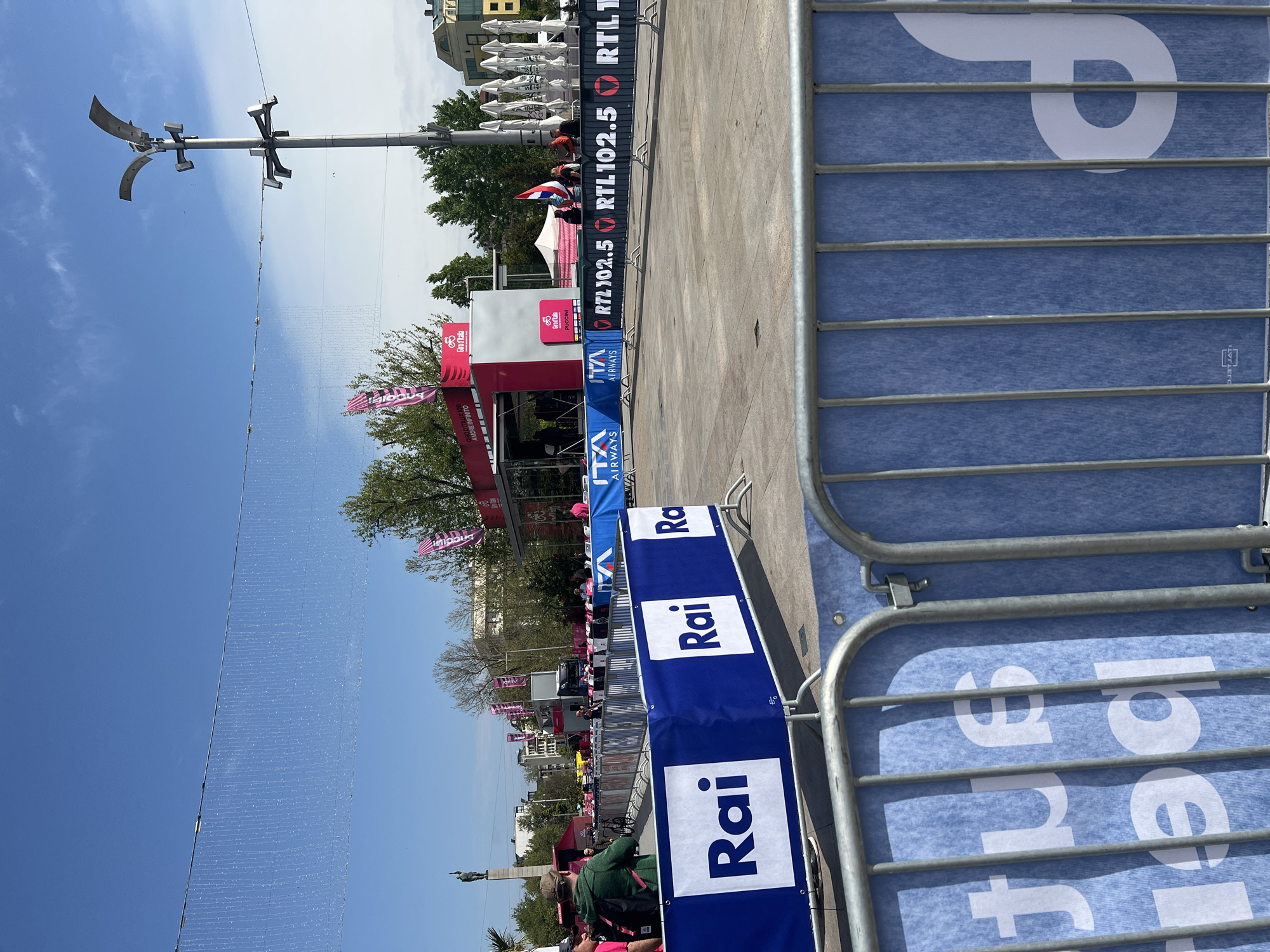 The image shows a street event, likely a sporting event, with branding for 'Rai', 'RTL 102.5', and 'Giro d'Italia'. There are metal barriers with advertisements. A stage or booth is visible in the background, decorated with flags and advertising ITA Airways. People are present but not clearly distinguishable. A distinctive street lamp is visible. The location is Burgas, Bulgaria.