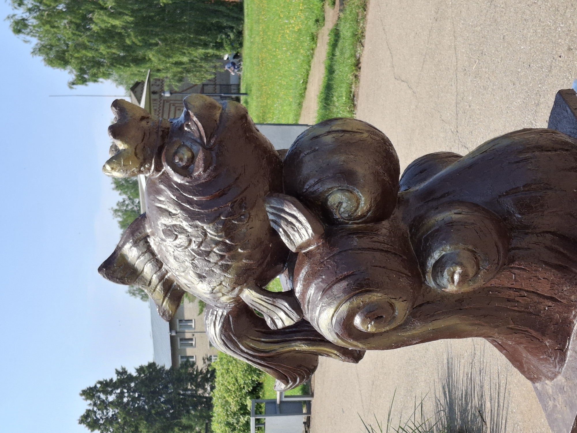 The image shows a brown, metallic sculpture of a crowned fish resting on what appears to be stylized waves. The sculpture is located in an outdoor setting with a paved walkway in the foreground and a grassy area with scattered yellow flowers in the background. Trees and a building are visible behind the grassy area, and a person on a bicycle can be seen in the distance. The overall scene suggests a public park or outdoor space.