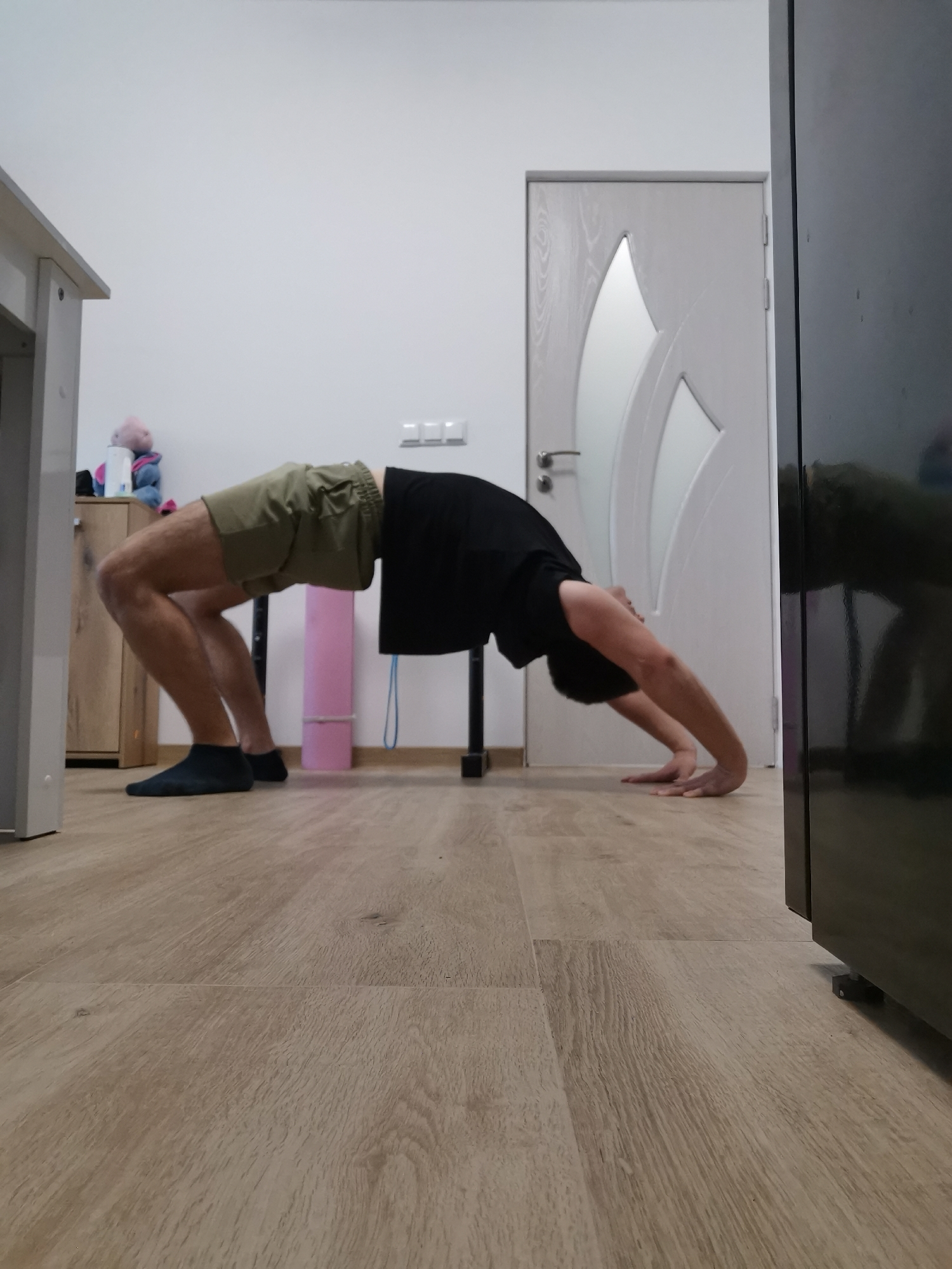 The image shows a person in a backbend position, likely a yoga pose, in an indoor setting. The person is wearing dark socks, khaki shorts, and a black t-shirt. Their hands are on the floor in front of them. Behind the person is a table and a cabinet with a stuffed animal and other objects on top. A closed white door with decorative inlays is visible in the background, along with electrical outlets on the wall. To the right of the door is a dark-colored cabinet or refrigerator. The floor is made of light-colored wood planks. There is no visual information to suggest the photo was taken in Burgas, Bulgaria. There are no specific clues about the location beyond it being a domestic interior.