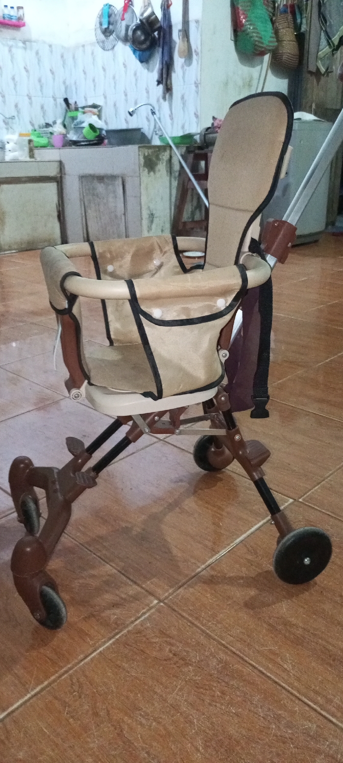 The image displays an unoccupied, tan and dark brown compact baby stroller situated on a light brown tiled floor within an indoor residential environment in Mns Alue, Indonesia. The stroller features a beige fabric seat with black trim and white button closures, supported by a dark brown and black metal frame with multiple small wheels. In the background, a kitchen area is visible, including a light-colored tiled countertop with a metallic faucet extending over a sink. Assorted kitchen items such as dishes, green plastic containers, and a white cat or object are present on the counter. The wall above the counter is covered with light-patterned tiles, from which various kitchen utensils, pots, a small fan, and woven baskets are suspended. To the right, a white appliance and a discolored light-colored wall are partially discernible. No individuals are present, and no actions are being performed.