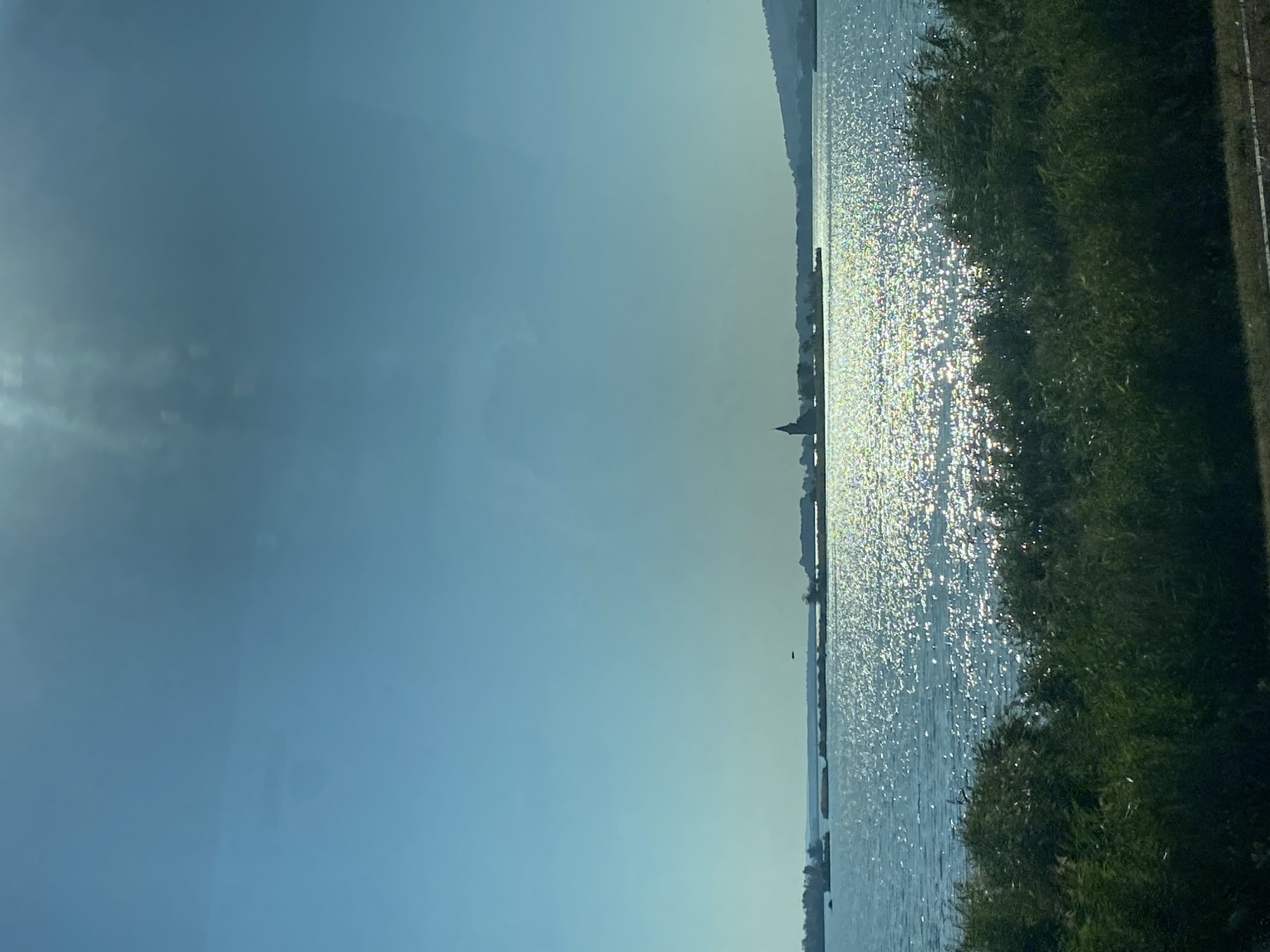 The image depicts a serene body of water with a distant silhouette of a building with a spire, likely a church, on a small island or peninsula. The water is shimmering with sunlight, creating a sparkling effect. In the foreground, there are lush green bushes and reeds. The sky is a clear, pale blue with some faint lens flare. The scene suggests a tranquil, natural landscape. This is likely the Pasohlávky Reservoir, also known as the Nové Mlýny Reservoir, in the South Moravian Region of the Czech Republic, near the town of Pasohlávky. The building with the spire is likely the Church of St. Linhart in Mušov, which was relocated due to the construction of the reservoir.