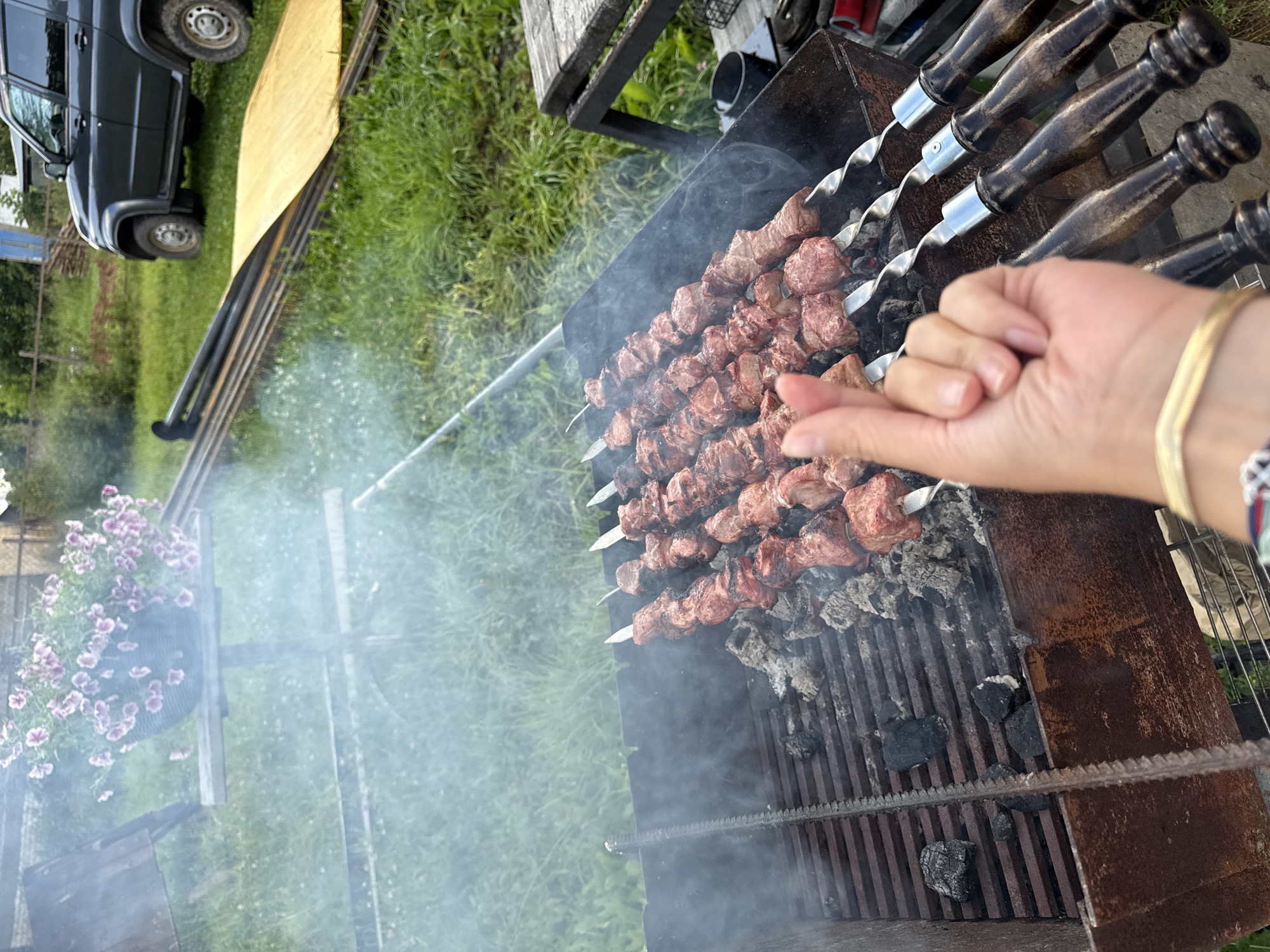 A person's hand is held up, making a heart shape gesture with their thumb and index finger, in front of a barbecue grill with several skewers of meat cooking over charcoal. The barbecue is outdoors, with green grass and some wooden structures in the background. A dark-colored SUV is parked to the right of the image. Smoke is rising from the grill. The overall scene suggests a casual outdoor gathering or meal preparation. The location is specified as Ветлужский, Russia.
