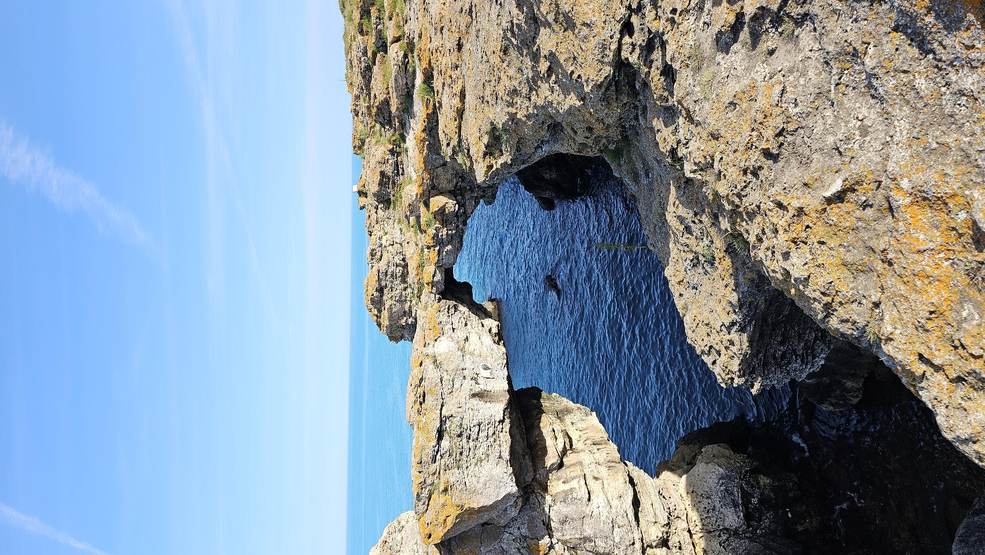 The image captures a coastal rock formation with a natural archway carved by the sea. The water within the arch is deep blue, contrasting with the light blue sky and sea visible on the left. The rocks are textured and colored in shades of brown, gray, and orange, with patches of green vegetation. The scene is oriented vertically, emphasizing the height of the rock structure. There are no people, specific subjects, distinct actions, or interactions visible in the frame. The environment is natural, marked by rock, sea, and sky. The location is unknown.