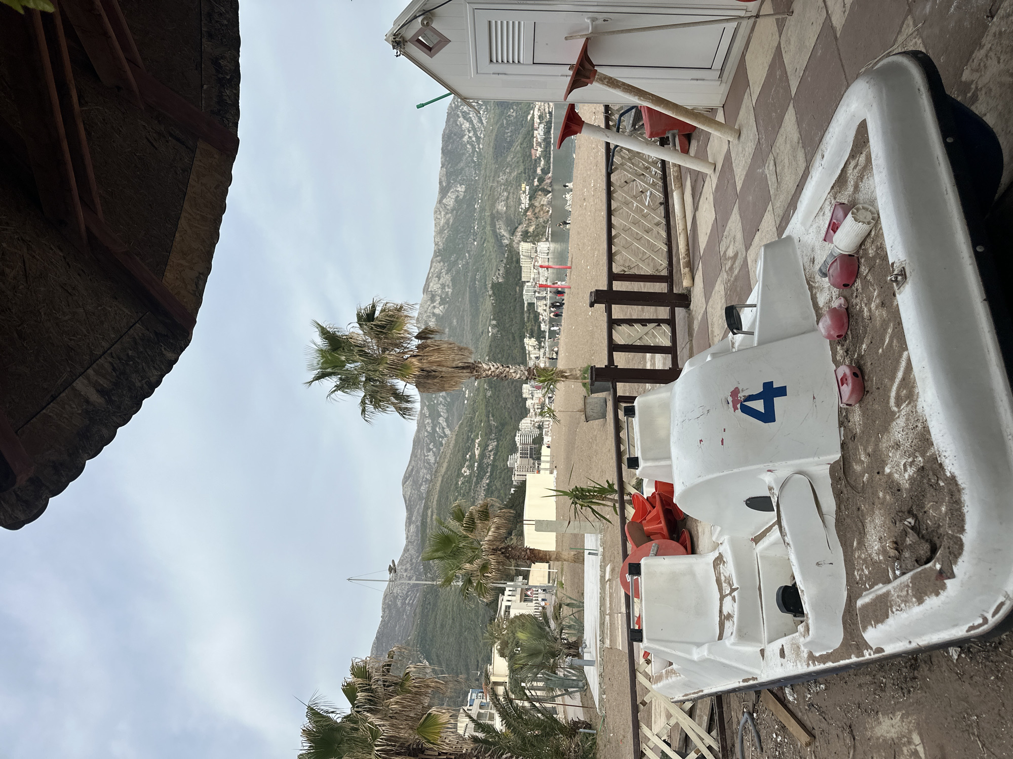 The image shows a beach scene in Boreti, Montenegro. A white pedal boat, labeled with the number "4," sits on a checkered tile surface, likely a patio or walkway adjacent to the beach. In the background, a sandy beach stretches out, backed by buildings and a tree-covered mountain. Palm trees are visible in the foreground and middle ground, adding to the beach atmosphere. A small white structure with a green accent is also present near the pedal boat, potentially a changing room or storage unit. The sky is overcast.