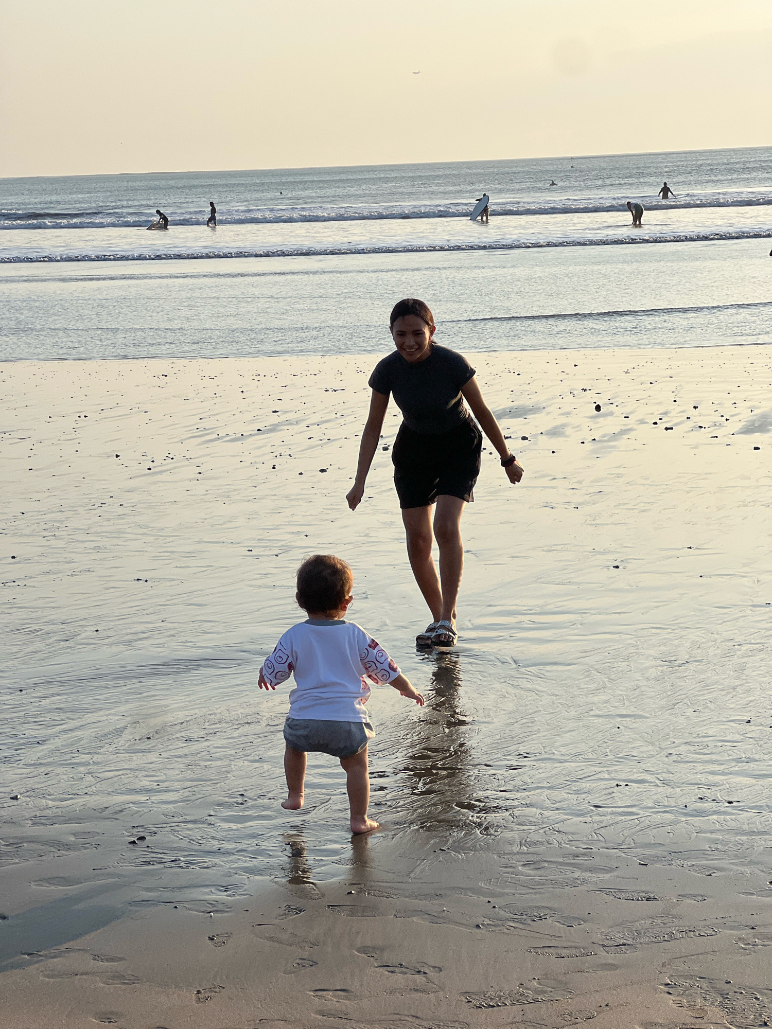 In Kuta, Indonesia, a mother and her young child are playing on the wet sand of a beach during sunset. The mother, wearing a dark t-shirt and shorts, stands with her arms outstretched, looking at her child. The child, facing away from the camera, is dressed in a white shirt with pink and purple designs and grey shorts. They are positioned a few feet apart, with the child in the foreground and the mother behind them. The shallow water laps at the shore, reflecting the warm, soft light of the setting sun. In the background, several other people are visible in the ocean, some with surfboards, enjoying the waves. The sand is marked with the footprints of those who have walked on it, creating a textured surface. The overall scene conveys a sense of joy and playful interaction between the mother and child in a serene beach environment.