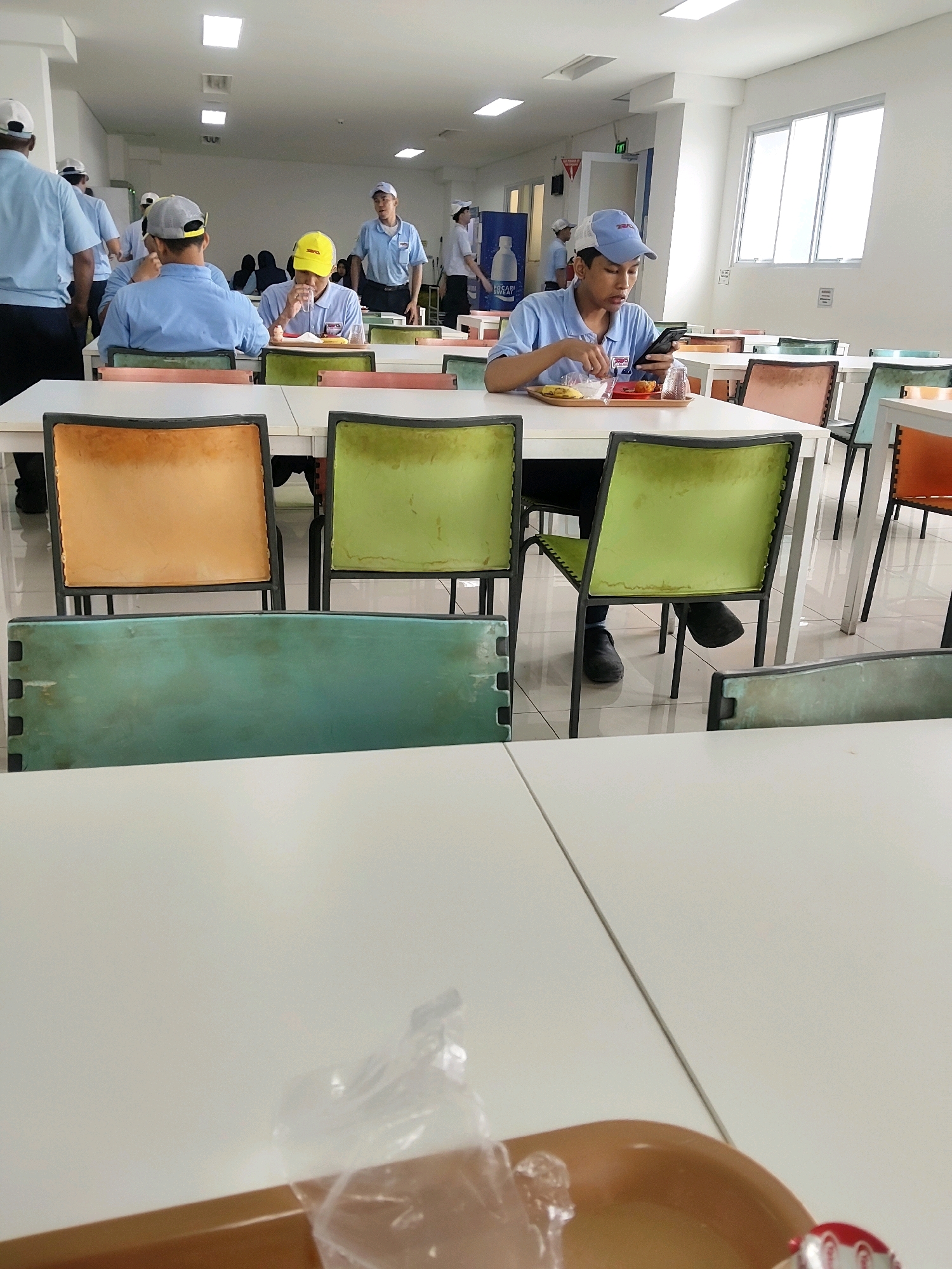 The image depicts an indoor cafeteria scene, likely within an industrial facility, given the attire of several individuals who are wearing light blue shirts and caps. The cafeteria features rows of tables with white laminate tops and dark metal legs. The chairs have colorful, cushioned seats in shades of orange, green, and turquoise. Fluorescent lighting panels are visible on the white ceiling.
In the foreground, a person wearing a blue shirt and a blue and white cap is seated at a table, looking down at a smartphone held in their left hand and holding a fork in their right hand. They appear to be eating from a tray containing food, including what looks like rice and a banana. A plastic cup is also on the tray.
In the middle ground, several other individuals are visible. One person in a yellow hard hat is seated at a table, looking down at their tray, also appearing to be eating. Another person in a blue shirt and grey cap is walking away from the camera. Further back, other people are seated or standing, some wearing similar uniforms.
The environment appears clean and functional, typical of a workplace cafeteria. The overall impression is of a break period where employees are having lunch and some are using their mobile phones. The location is stated to be in Mekarmukti, Indonesia.