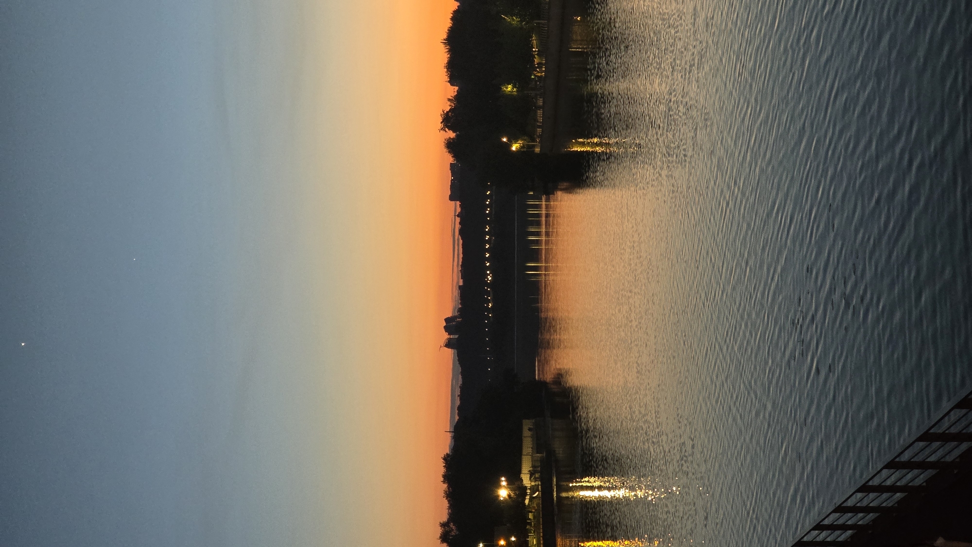 The image captures a sunset over the Dnipro River in Kyiv, Ukraine. The sky transitions from a deep blue at the top to a vibrant orange and yellow near the horizon, with two faint stars visible in the upper portion. Silhouetted trees line the riverbanks, and streetlights glow along the distant bridge, creating reflections on the water's surface. In the foreground, a dark metal railing runs along the edge of a walkway. The water itself is calm, with gentle ripples disturbing the reflections of the lights and the sky. The overall scene evokes a peaceful evening atmosphere.
