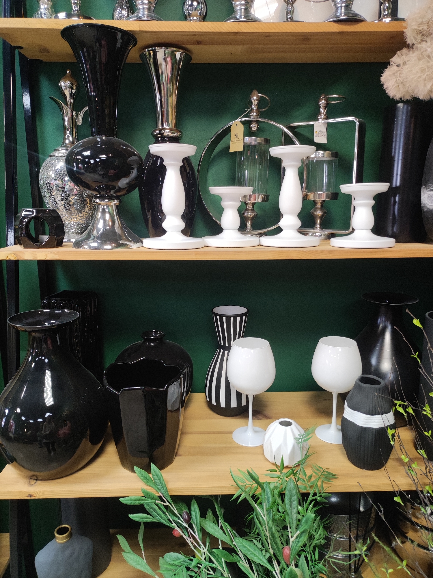 The image shows a shelf display of decorative vases and candle holders, predominantly in black, white, and silver. The top shelf features an assortment of black vases, silver candle holders, and white pedestals. The bottom shelf displays various black vases of different shapes and sizes, two white wine glasses, and two small black and white decorative objects, alongside artificial greenery with olives. The background is a solid dark green. The location and presence of people cannot be determined from this image. It's not possible to confirm if this display is located in the city of Votkinsk, Russia based on visual evidence alone.