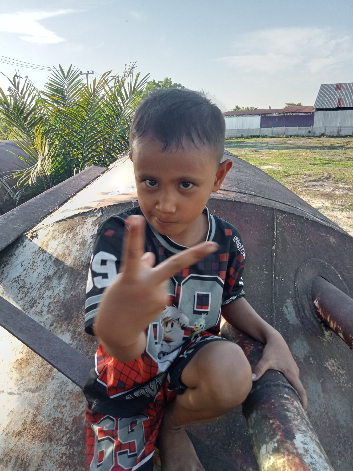 A young boy with dark hair and brown eyes, wearing a black, red, and white patterned t-shirt and shorts, kneels on a large, rusty, cylindrical object. His right hand is raised, with his index and middle fingers extended, forming a "peace" sign. The boy is positioned in the center of the frame, facing forward.
The object he is kneeling on appears to be industrial or salvaged material, showing significant signs of rust and wear. It has a curved surface and at least one pipe extending from it.
The background suggests an outdoor, semi-rural or industrial setting. To the left, there are palm fronds and utility poles with wires. In the distance, several buildings with corrugated metal roofs are visible, with a fence in front of them. The ground is covered in dry grass and some patchy dirt. The sky is clear with a few wispy clouds, and the lighting indicates it is daytime, likely late afternoon due to the warm light. The overall impression is a candid, outdoor portrait of a child in a somewhat neglected or industrial area of Kualu, Indonesia.