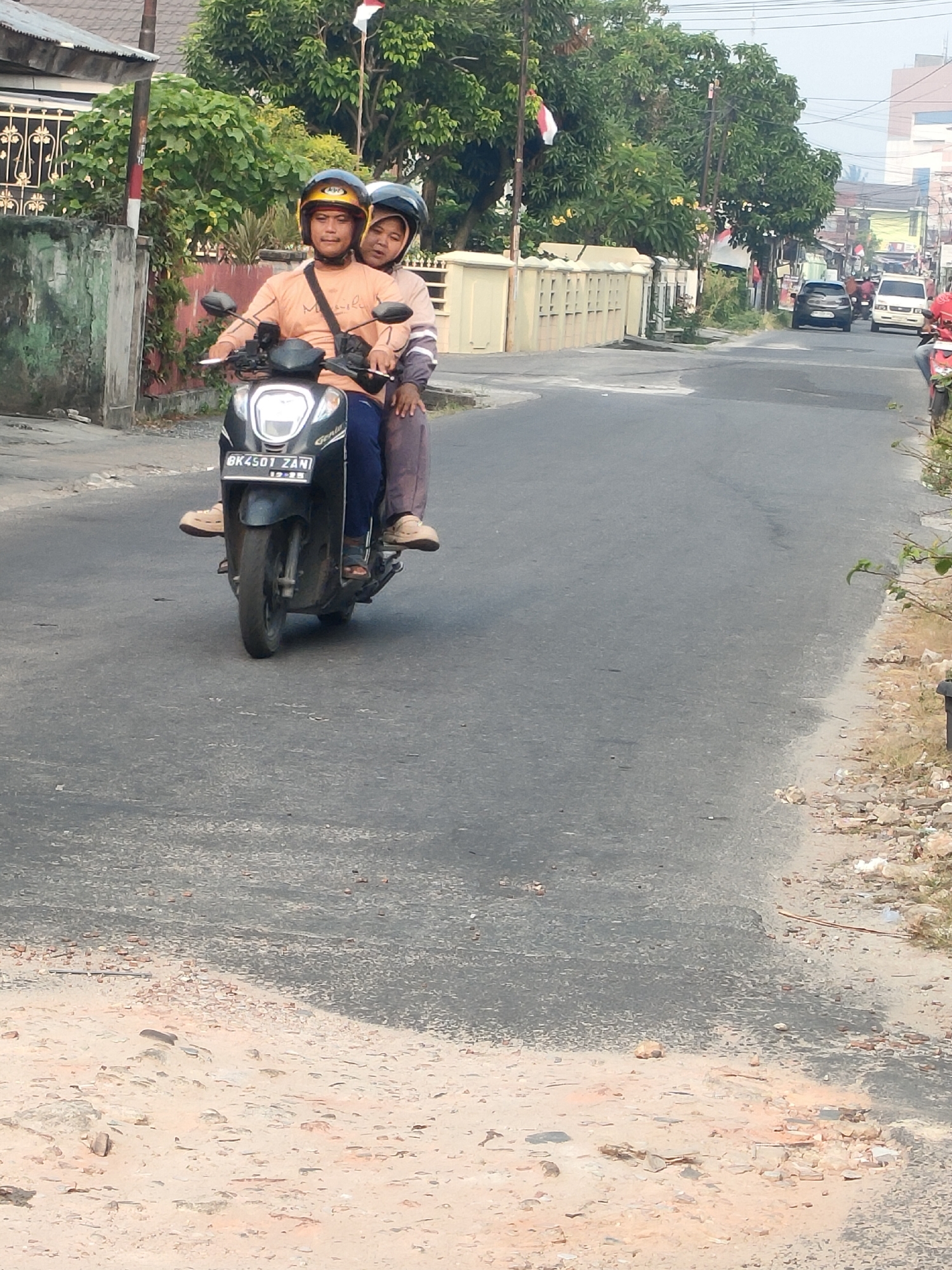 Two people are riding a black scooter on a paved road. The person in front, wearing an orange shirt and a yellow helmet, is driving. The person behind, wearing a light pink top and a dark helmet, is a passenger. The scooter has a license plate that reads "BK 4501 ZAN" and "12-25". They are on a street lined with buildings and trees, with some Indonesian flags visible. The road surface is asphalt, with a sandy and rocky shoulder on the right side. In the background, several vehicles are driving on the road, including a black car and two white vehicles. The scene appears to be in Pekanbaru, Indonesia, based on the license plate format which is common in the Riau province.