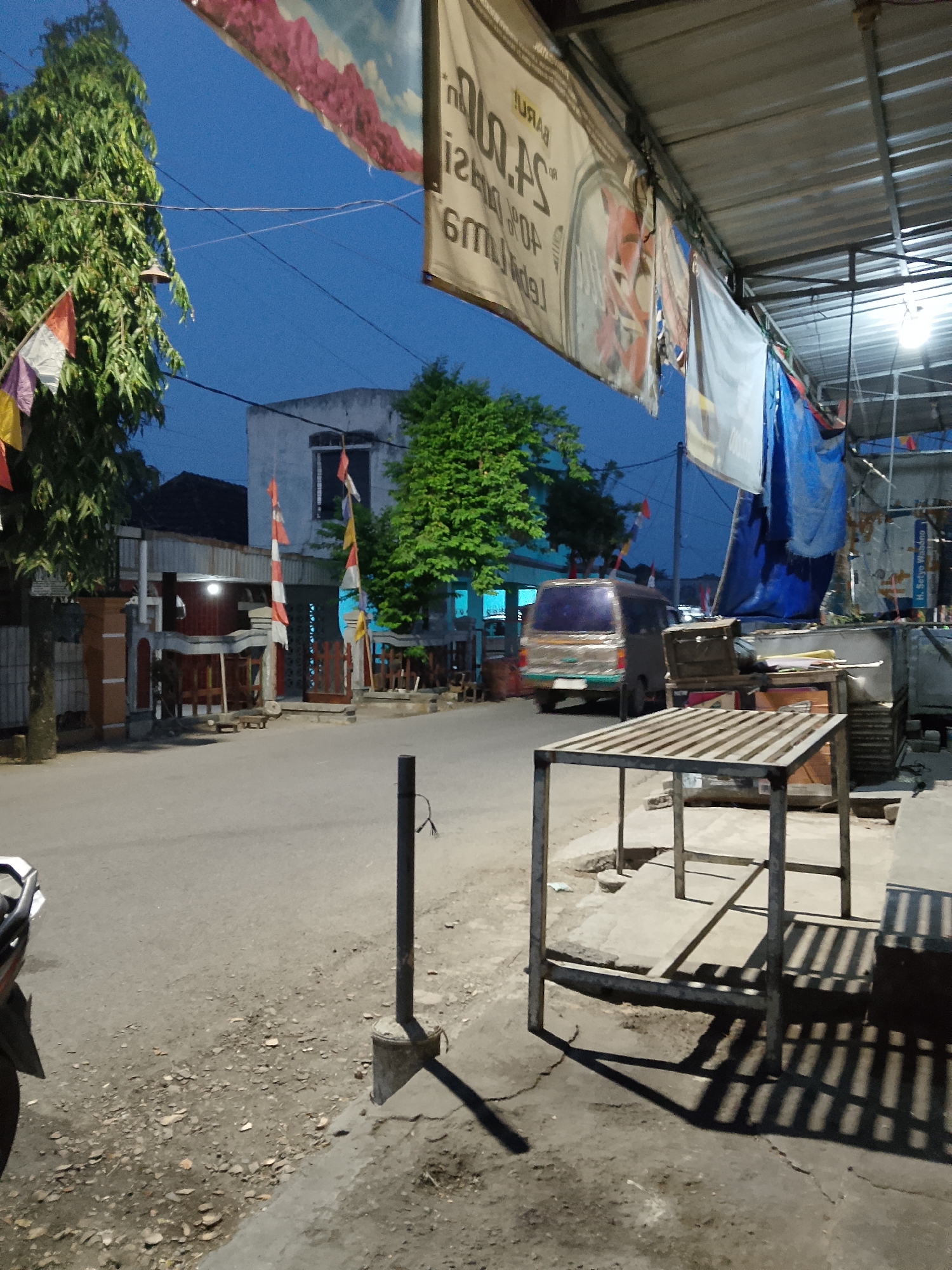 The image depicts a street scene in the city of Padangan, Indonesia, during twilight hours. The foreground features a metal table with a slatted surface, casting distinct shadows on the concrete ground. To the left, the rear of a motorcycle is visible. A dark pole with exposed wires stands near the table. The street itself is paved and appears somewhat worn, with scattered debris.
Further back, a silver van is parked on the side of the road, facing away from the camera. Adjacent to the road, there are buildings, one of which has a white facade and a dark window. Red and white flags are displayed prominently in front of this building. Lush green trees are scattered throughout the scene, adding to the urban landscape. Above, several banners hang from a covered structure, some displaying text and graphics. The sky is a deep blue, indicating the fading light of dusk. The overall impression is of a quiet street corner in a local Indonesian town as evening sets in.