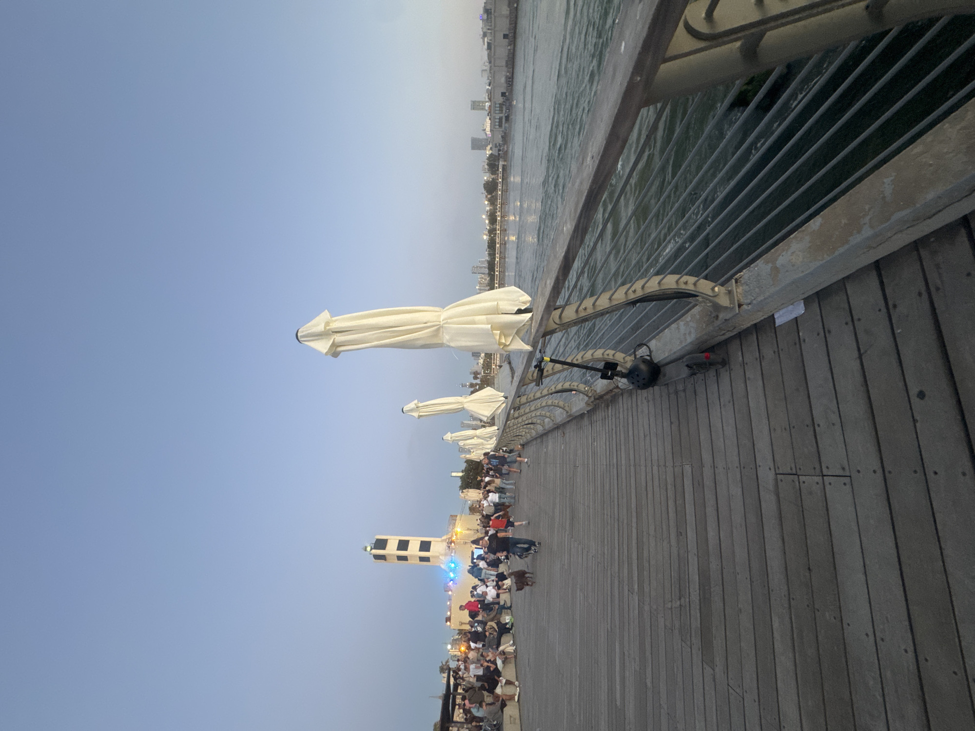 The image captures a scene along a boardwalk in Tel Aviv, Israel. The boardwalk, constructed of wooden planks, runs parallel to the Mediterranean Sea, which is visible on the right side of the frame. A metal railing separates the boardwalk from the sea. Several closed, white parasols line the boardwalk. Numerous people are present, some walking and others gathered in groups. In the distance, a white tower stands prominently. The sky is clear. The overall impression is of an evening stroll along a popular Tel Aviv beachfront location.