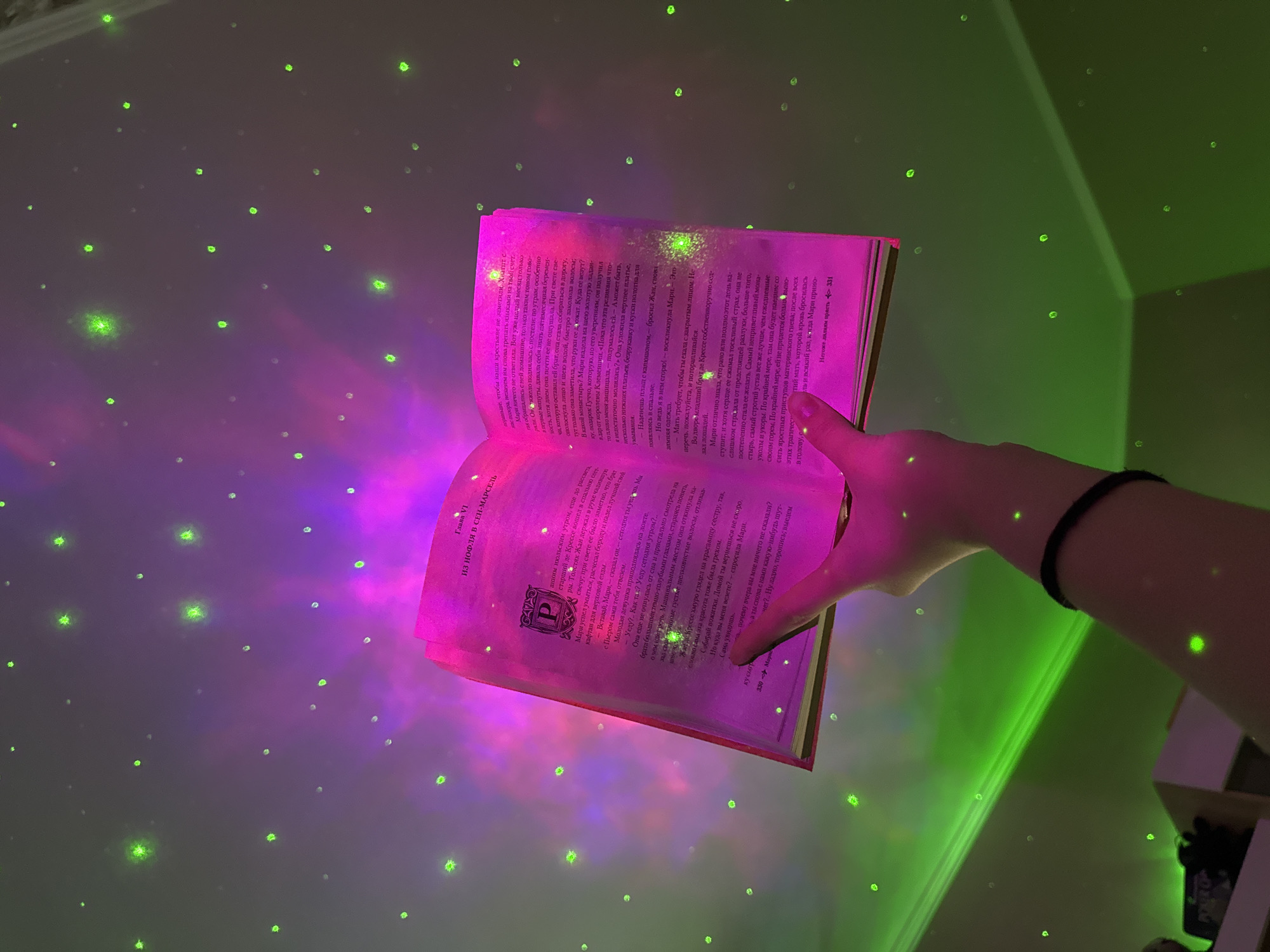 A person's arm and hand hold an open book illuminated with pink light against a backdrop of green and purple light patterns on a wall. The book's pages are filled with text in a Cyrillic script. The person wears a black wristband. The light patterns consist of scattered green dots and larger areas of pink and green gradients. A shelf with visible objects is in the lower right corner. The context suggests the interior of a room.