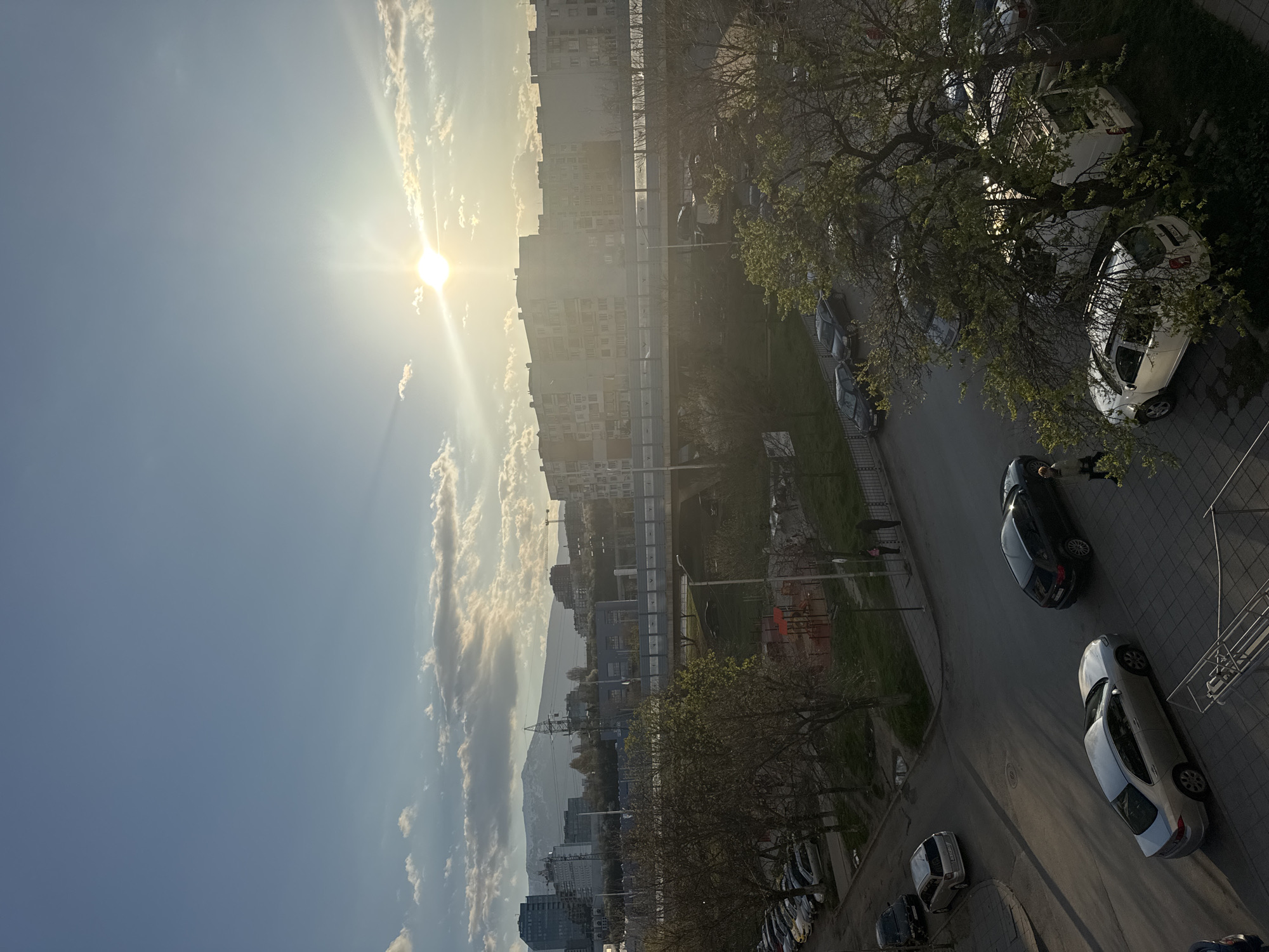 The image depicts an elevated view of a street scene in Sofia, Bulgaria. The sun shines brightly over the cityscape, partially obscured by light clouds. Apartment buildings of varying heights line the horizon, with a mountain range visible in the distance. A street runs through the foreground, lined with parked cars and trees beginning to sprout leaves. A person is seen standing near a black car parked on the side of the street. A green space with some construction or renovation appears between the street and the buildings. Power lines are visible.