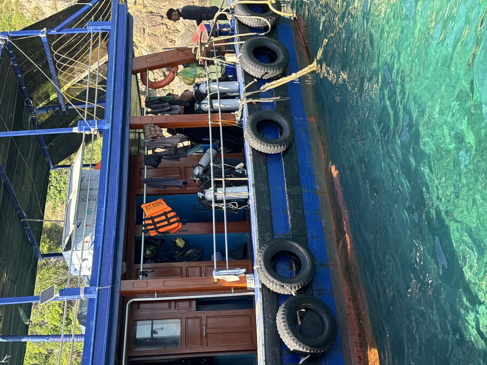 The image shows the side of a blue and brown boat docked in clear, turquoise water. The boat is laden with diving equipment, including several scuba tanks, a life vest, and diving masks. Clothing hangs from a railing on the side of the boat, suggesting recent activity or preparation for a dive. A person in dark clothing stands on the deck near the stern of the boat, looking out towards the water. The background consists of a rocky, green cliff face. The scene appears to be a typical view of a dive boat operation in the coastal waters near Nha Trang, Vietnam, known for its coral reefs and diving excursions.