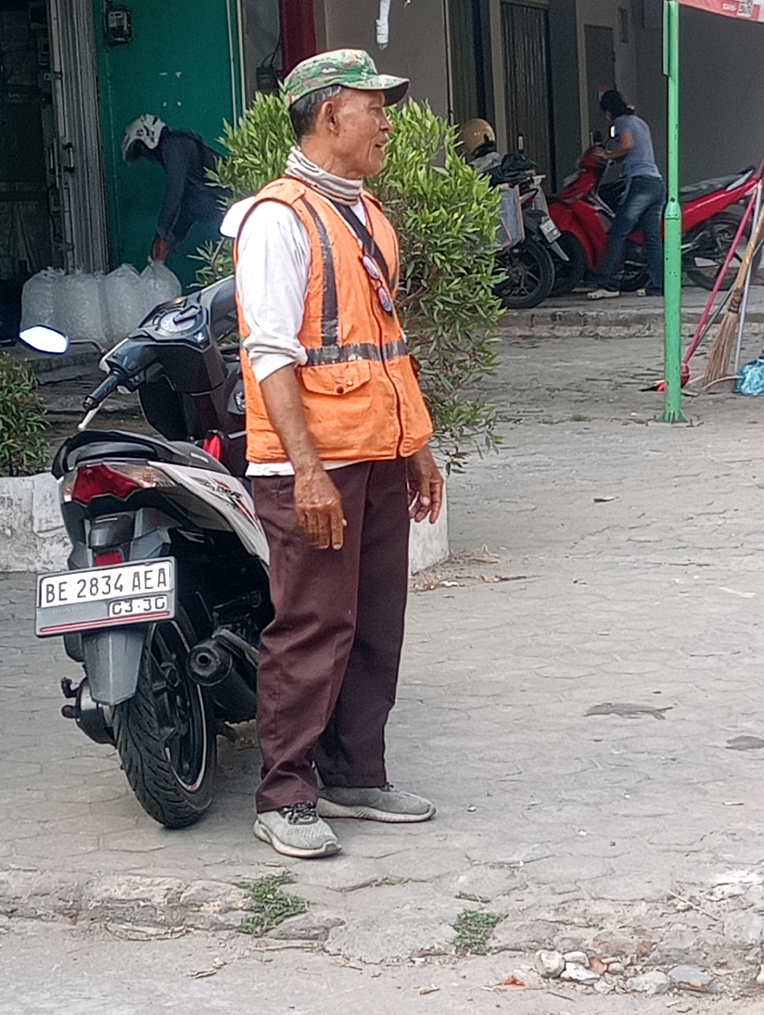 An outdoor, daytime, medium shot captures a man wearing an orange safety vest over a white shirt and brown pants, standing beside a motorcycle. He has a camouflage baseball cap on and appears to be looking to his left. A motorcycle with a license plate reading "BE 2834 AEA" is parked on a paved surface in the foreground. In the background, another person in dark clothing is visible near a building, and a red motorcycle is parked to the right. The scene appears to be in Bandar Lampung, Indonesia.