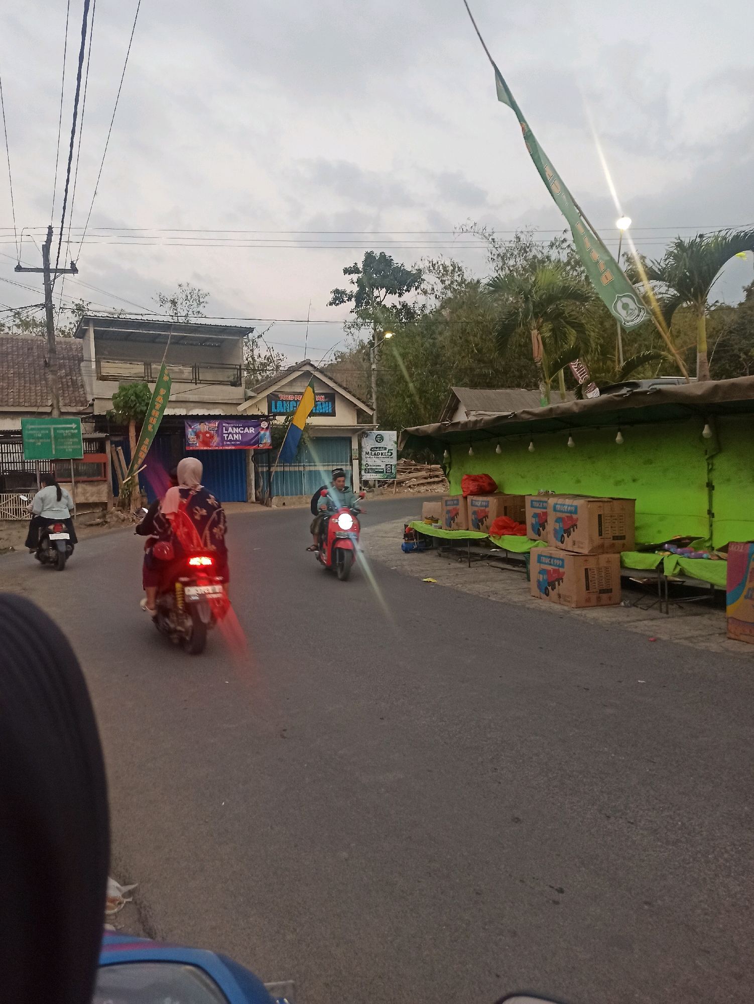 The image captures a street scene in Tlogosari, Indonesia, likely during late afternoon or early evening. The foreground is dominated by the dark asphalt road, with a blue scooter partially visible at the very bottom. Further down the road, two individuals on a red motorcycle are riding away from the camera, the rear brake light emitting a distinct red glow. To the left, another person is riding a white scooter. On the right side of the road, a green awning covers a market stall displaying several cardboard boxes with images of trucks and the text "TRUCK 999". Red fabric is also visible on the stall. In the background, buildings with various roofs and facades line the street, some adorned with flags, including green banners with logos and text. Palm trees and other greenery are interspersed between the structures. Utility poles and overhead wires are present, indicating an urban or semi-urban setting. The sky is overcast, suggesting a cloudy day. The overall impression is one of everyday life in a bustling local area, with commerce and transportation visible.