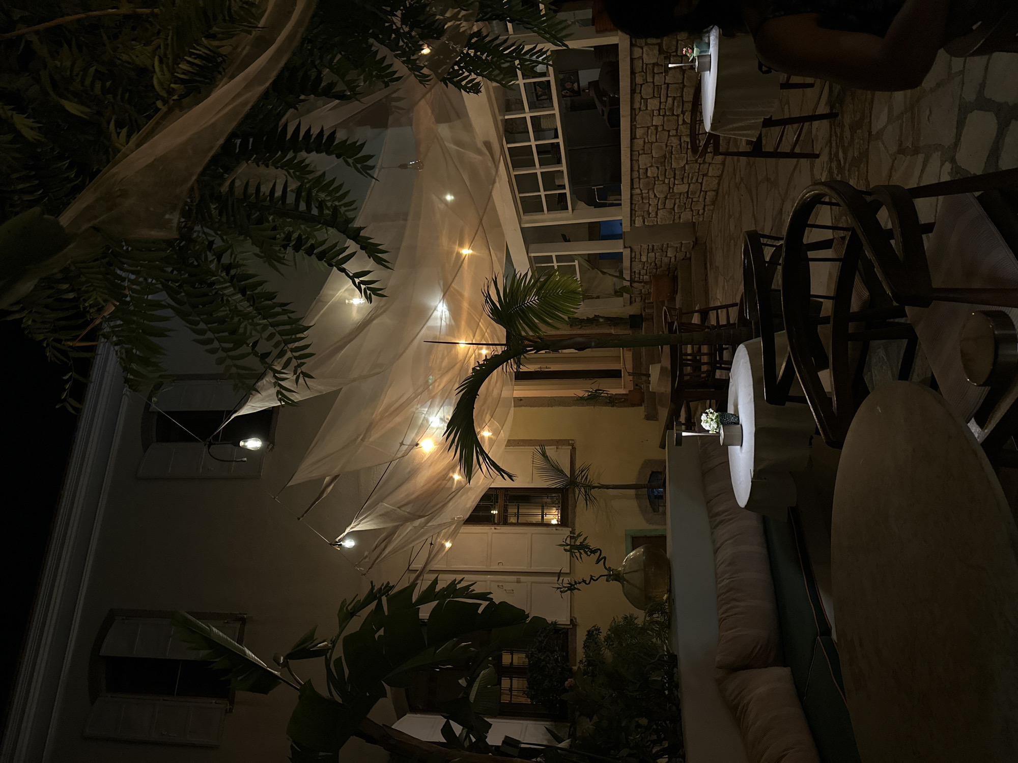 This image depicts an outdoor seating area at night, likely a restaurant or cafe, in the city of Urla, Turkey. The scene is lit by string lights draped across a sheer fabric canopy overhead and by small table lamps.
In the foreground, two round tables are visible. The closest one is large and indistinct due to low light. The table to the right is smaller, covered with a white cloth, and has a small lamp and a floral arrangement on it. Around the tables, several dark wooden chairs are arranged.
To the left, a long, cushioned bench seating area is present, with pillows. Behind the bench, a large glass vase with a curved neck sits on a ledge, holding a delicate plant. Lush green plants, including palm-like foliage, are scattered throughout the scene, adding to the ambiance.
The background features a building with yellow walls and windows with white shutters. One window has an open shutter, and a light bulb hangs from a cord just outside it. Another window shows a reflection of light. A stone wall with a rough texture forms part of the background structure.
The overall impression is a charming and atmospheric nighttime setting, typical of a Mediterranean outdoor dining experience.