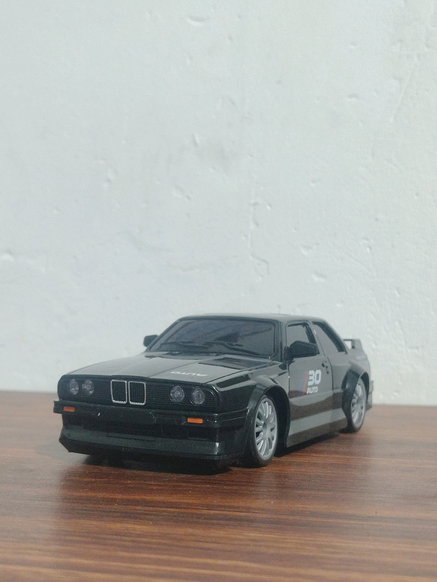 A black toy car, resembling a BMW E30 M3, is positioned on a wooden surface. The car faces forward and slightly to the left. It features silver rims and a decal on the side with the text "30 AUTO". The background is a textured, off-white wall. The scene is lit from the front, casting a slight shadow behind the car. There is no indication of the city of Trincomalee, Sri Lanka in the image.