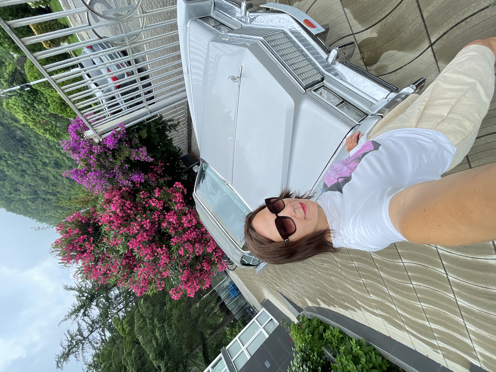 A woman in sunglasses and a white t-shirt takes a selfie in front of a vintage silver car. The car's hood and grill are visible. Behind the car, there's a large bush with pink and purple flowers and a metal fence. In the background, there are trees and a cloudy sky. To the left, part of a modern building with large windows is visible, and green bushes line the walkway. The ground is paved with interlocking stones that appear wet. The scene is likely outdoors in a temperate climate. The presence of a vintage car and the architecture suggest a location with a mix of historical and modern elements.