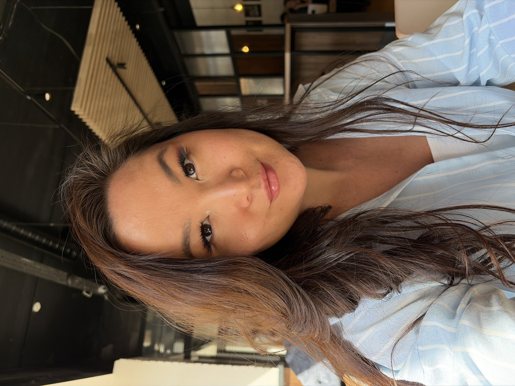 A woman with brown hair and fair skin is looking directly at the camera. She is wearing a light blue and white striped shirt and a white top underneath. Her makeup includes eyeshadow and mascara. The background appears to be an indoor setting, possibly a cafe or restaurant, with wooden ceiling panels, dark walls, and tables visible. There are no specific indicators of the city of Chaikovsky, Russia within the image.