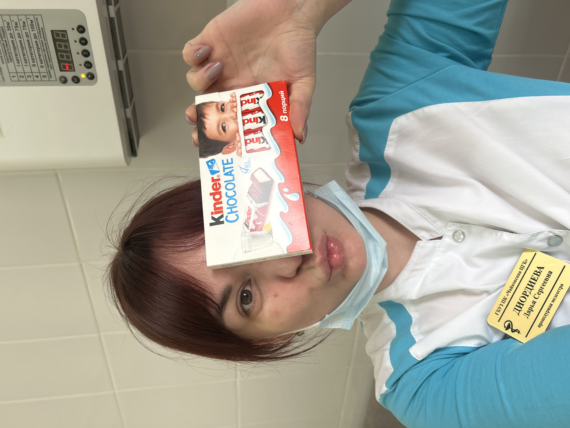 Here is a description of the image.
**Scene:** The image depicts a person in a medical setting, possibly a hospital or clinic, with a box of Kinder Chocolate positioned over their forehead. The person, identified as Darya Sergeevna Diordieva, a procedural nurse from the Chaykovskaya Central City Hospital, is wearing a medical uniform with a name badge. They also have on a surgical mask which is pulled down from the nose area, and are making a "kissing" face expression.
**Environment:** The background suggests an indoor, clinical setting. The wall behind the person is tiled, and there is some type of air conditioning unit mounted on the wall above.
**Objects:**
* Kinder Chocolate box (labeled in Russian)
* Medical uniform
* Name badge
* Surgical mask
* Air conditioning unit
* Control panel
**Location:** Based on the name badge information and the OCR text, the image is likely taken in or near Chaykovsky, Russia, which is in the Perm Krai region of Russia.
**Summary:** The overall impression is a posed photo, with the nurse making a playful face with a box of chocolate.
