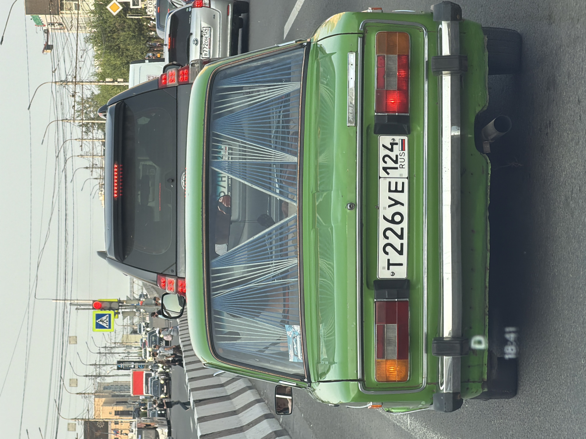 The image shows the rear of a bright green, older model car, possibly a Soviet-era Lada, driving in traffic in the city of Krasnoyarsk, Russia. The car has a license plate that reads "T226YE 124 RUS". The rear window is partially covered by decorative blue and white blinds. In the background, other vehicles are visible, including an SUV and a silver sedan. Traffic lights and street lamps are present, indicating an urban environment. A pedestrian crossing signal is visible in the distance, showing a red light. The overall scene depicts a typical city traffic situation.