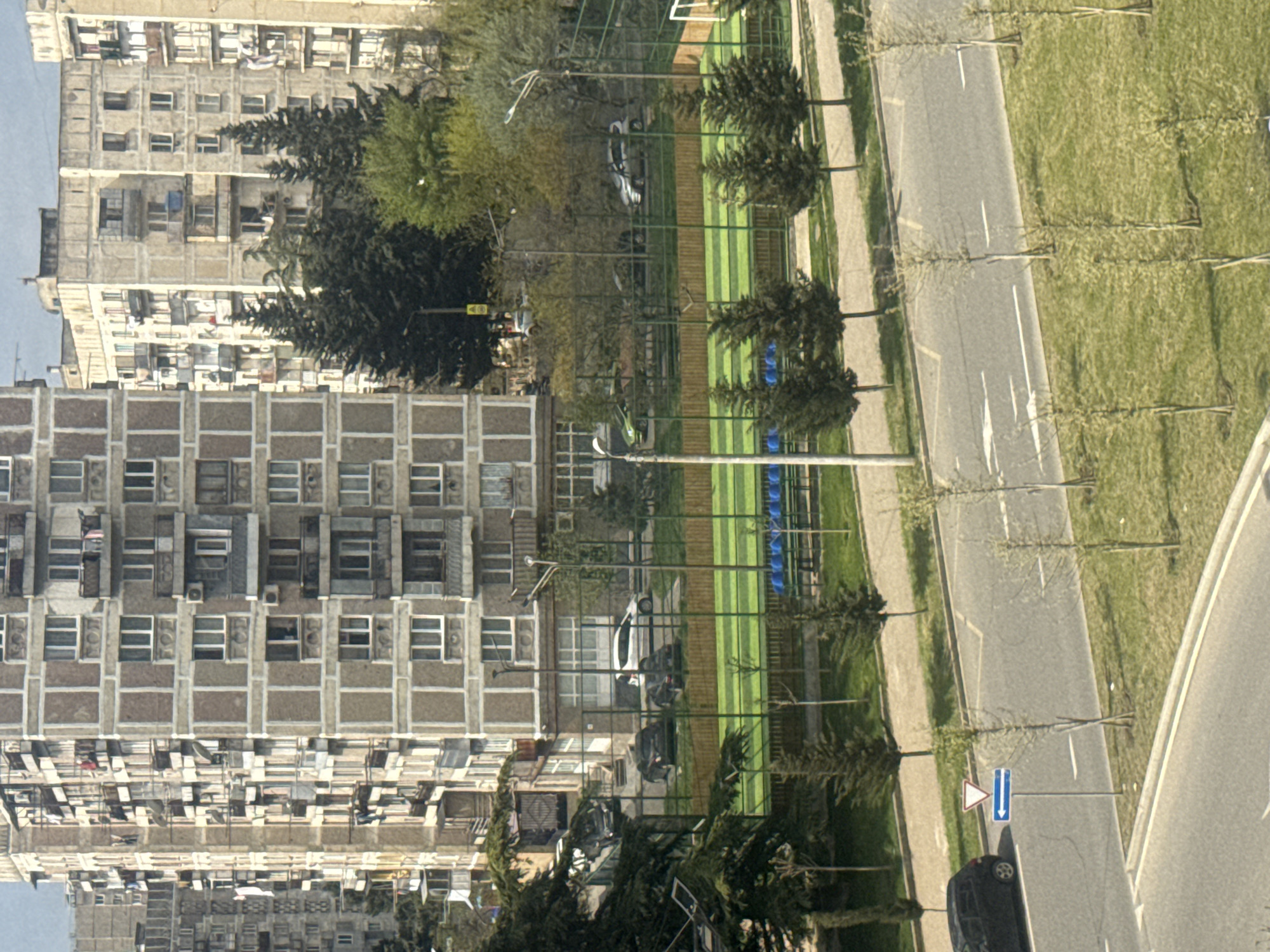 The image shows an urban scene in Tbilisi, Georgia. The architecture is dominated by Soviet-era apartment buildings, characterized by repetitive patterns of windows and balconies. Alongside these buildings, there's a sports complex with a green field and blue stadium seating visible through a fence. A road runs parallel to the complex, featuring road markings and a street sign. The environment includes greenery, with trees lining the road and scattered throughout the complex. Several cars are parked nearby. The scene appears to be shot from an elevated viewpoint.