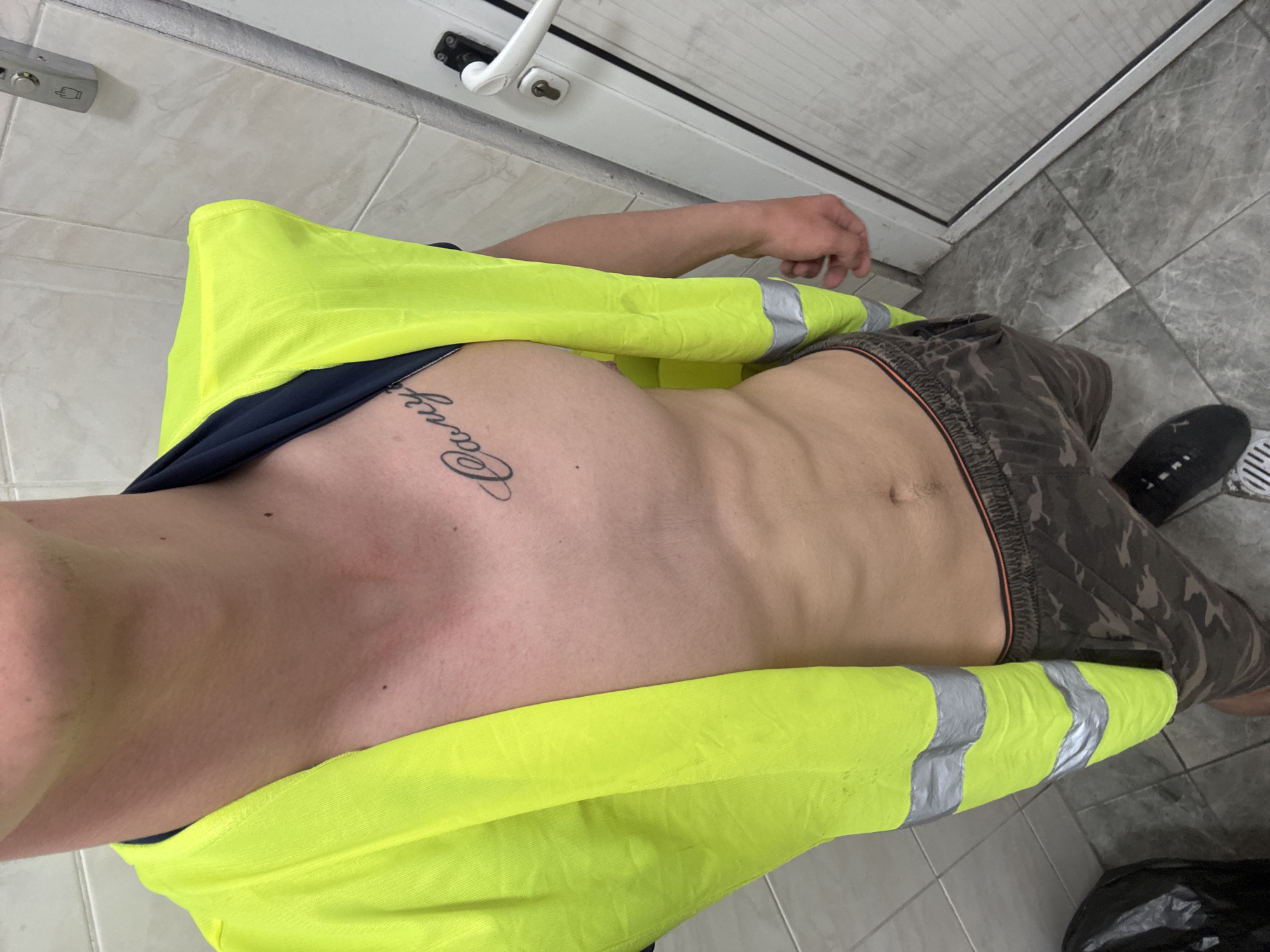 A top-down shot shows a person wearing a yellow safety vest, dark shorts with a camouflage pattern, and black shoes. The person has a tattoo on their upper chest. The environment includes light gray tile and a white door. The location is not discernible as Varna, Bulgaria.