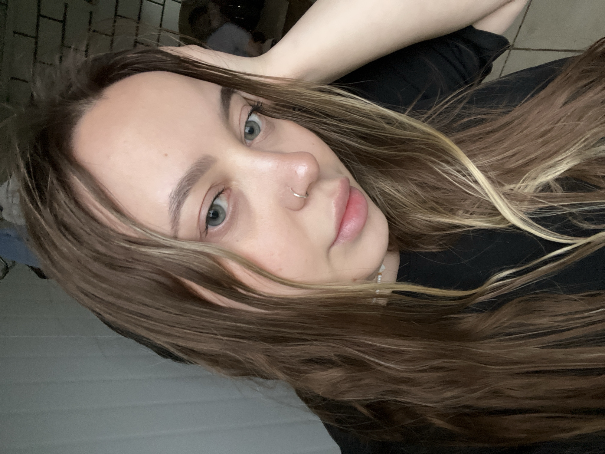 A selfie of a woman with long brown hair, styled with blonde highlights, is captured from a slightly high angle. She has fair skin, green eyes, and a nose piercing. She wears a black top. The background includes a tiled wall and the edge of a white wall. The location is not identifiable.