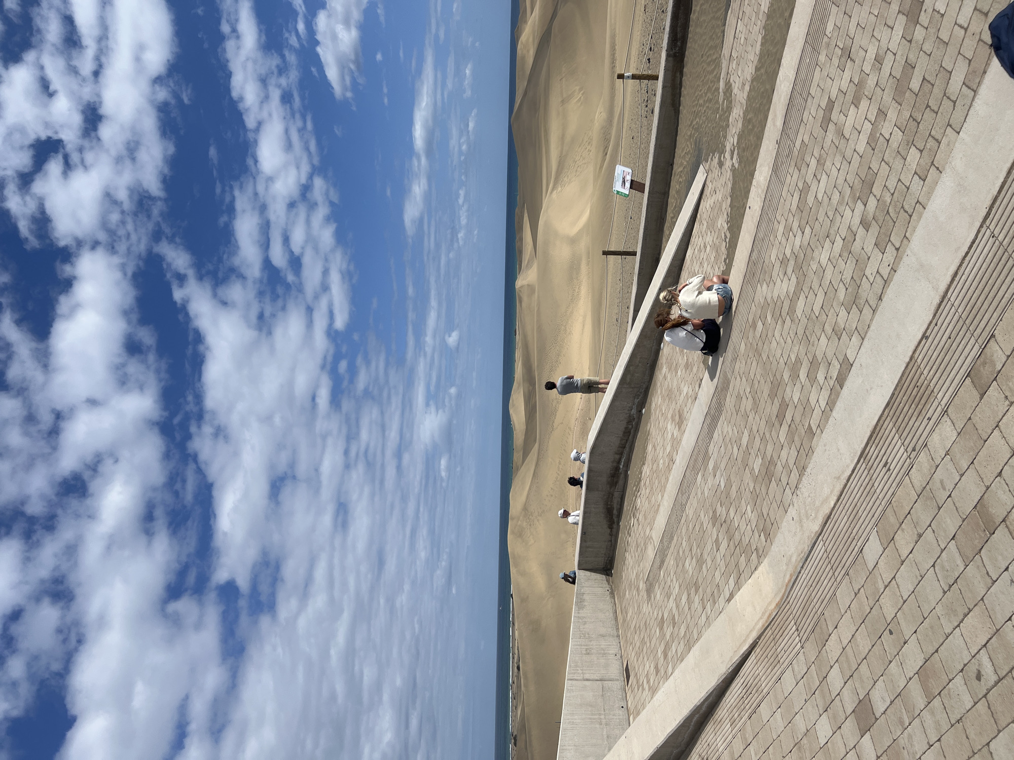 The image shows a scene in San Bartolomé de Tirajana, Spain, where sand dunes meet the ocean under a sky with patchy clouds. People are present at an elevated walkway and viewing area made of brick-like blocks and concrete, observing the dunes. A man stands on the walkway while two women are crouched down. Other people are also present, standing along the walkway. The landscape consists of sand dunes and the adjacent ocean stretching towards the horizon. The sky is predominantly blue with scattered white clouds.