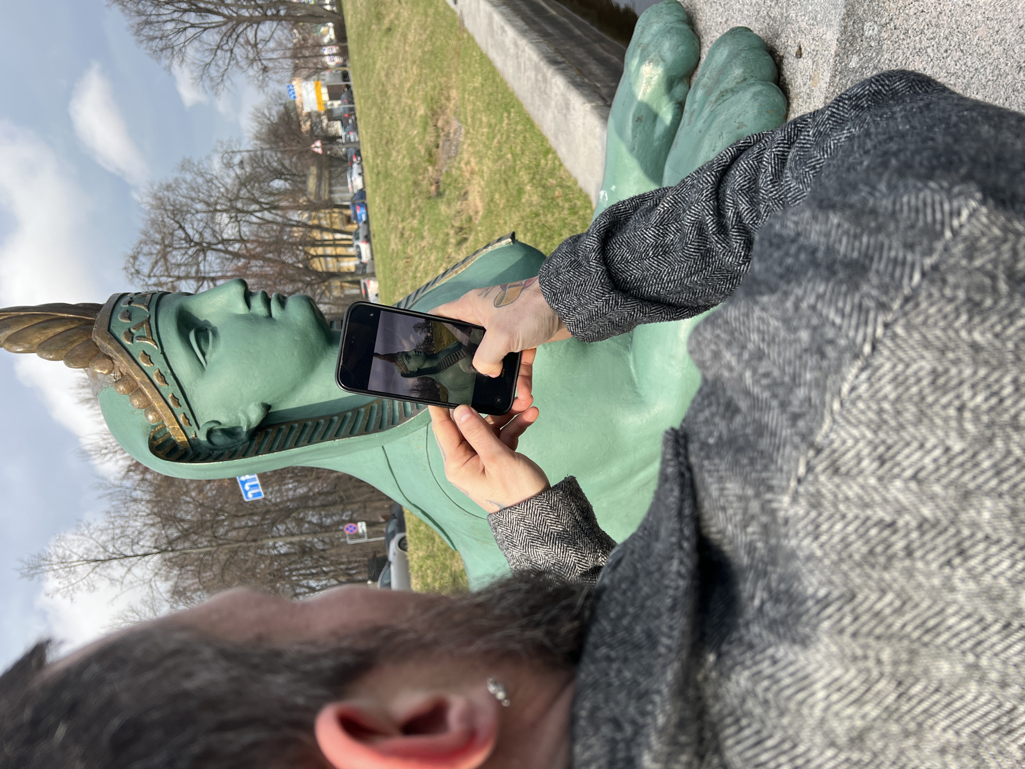 A man is taking a photo of a statue. The statue is of a sphinx-like figure. The background is a park-like setting, and there are buildings in the distance. The location is likely Saint Petersburg, Russia.