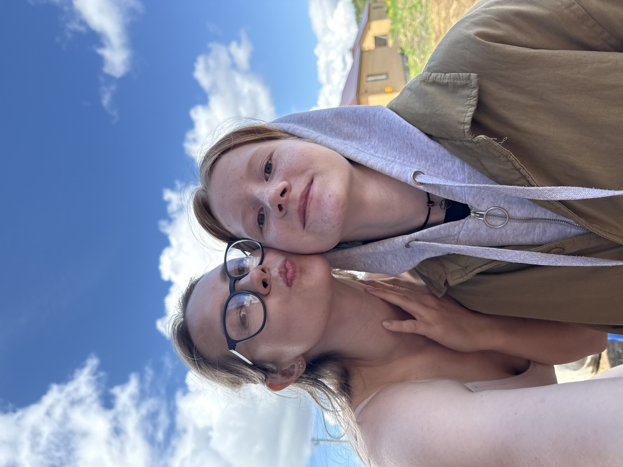 Two women are closely posing for a selfie with a blue sky filled with white clouds in the background. One woman is wearing glasses and making a kissing face while the other woman is wearing a hoodie and a jacket. The background shows a building and some vegetation. The location is in the city of Chaikovsky, Russia.