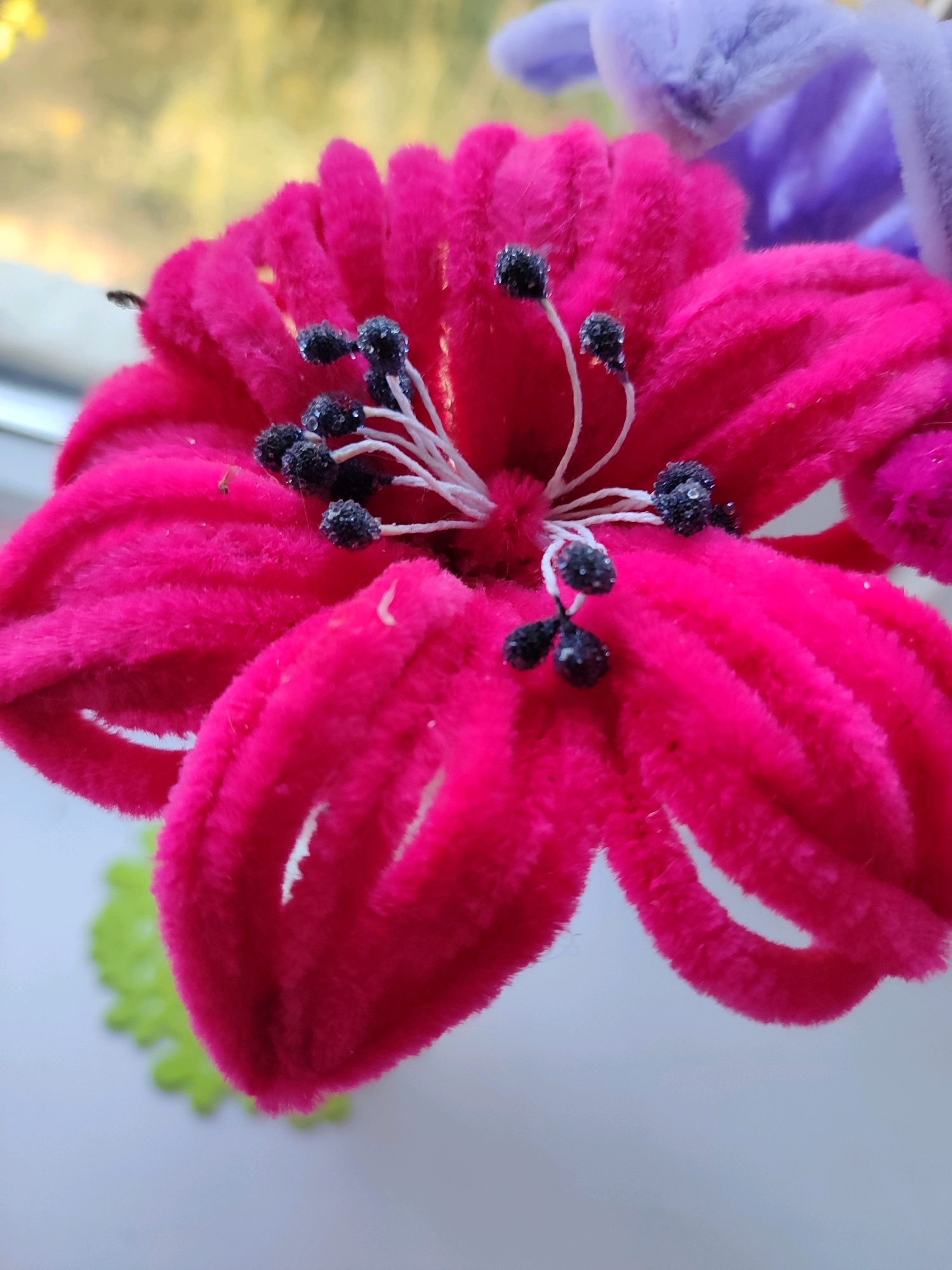 A close-up, slightly angled view shows a fluffy, bright pink pipe cleaner flower. The flower has five prominent petals that curve outwards and are covered in a soft, velvety texture. In the center of the flower are several thin white filaments tipped with small, dark, glittery beads resembling stamens. In the background, a blurry light purple pipe cleaner flower is visible, along with a soft yellow, out-of-focus background that suggests an outdoor setting or a window. A small, out-of-focus green object with serrated edges is visible in the lower left quadrant. The overall impression is of artificial flowers, likely part of a decoration or craft. There is no indication of a specific city or location beyond a general indoor or outdoor setting.