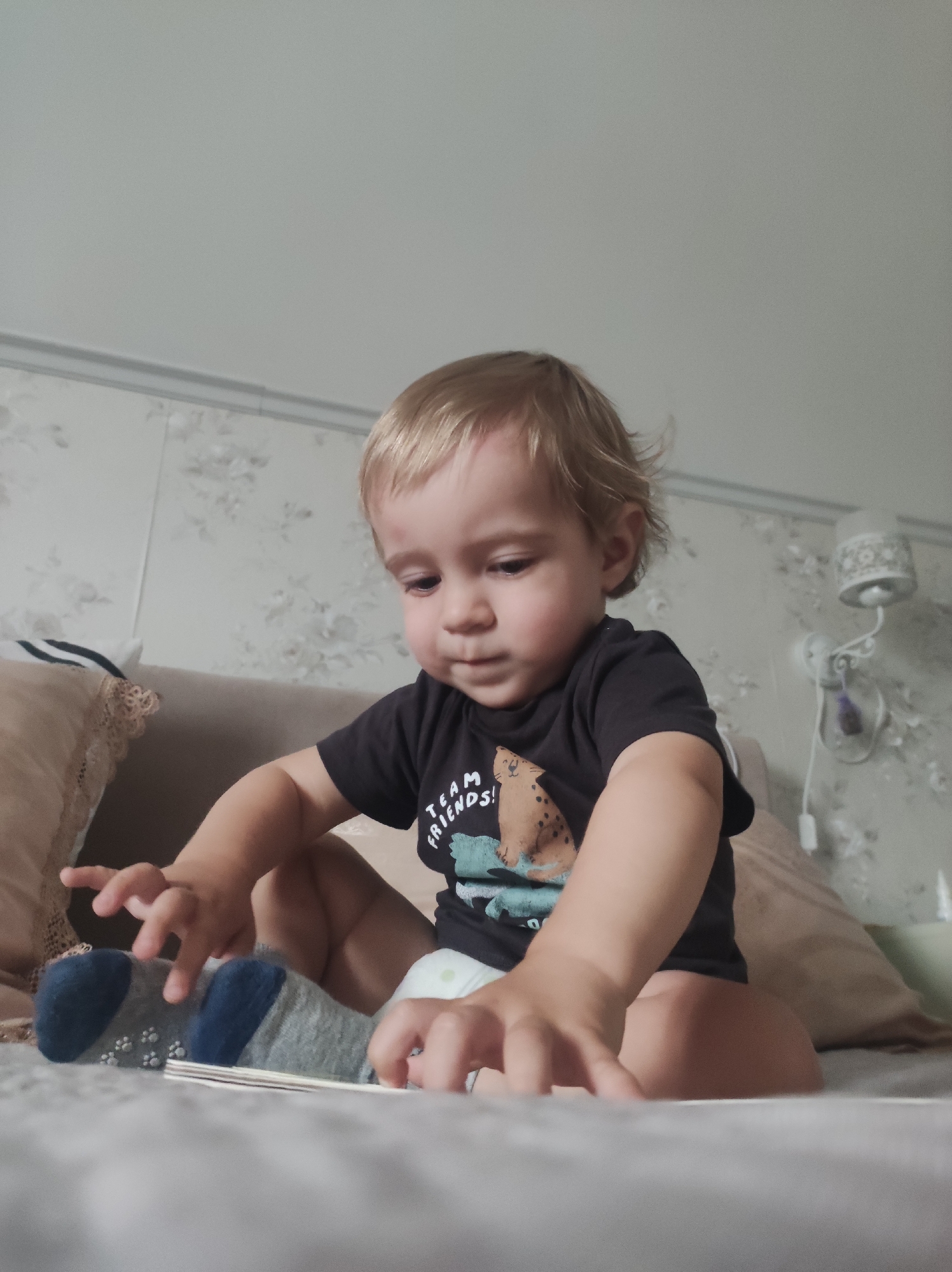 A young child, likely a toddler with blonde hair, is seated on a bed or sofa in an indoor setting. The child is wearing a dark t-shirt, a diaper, and gray and blue socks with white paw print anti-slip grips on the soles. The t-shirt features a graphic of a spotted animal, possibly a leopard, and partially visible text that reads "T_AM FRIENDS!", suggesting "TEAM FRIENDS!".
The child is looking downwards with a focused expression, their hands reaching out towards a flat object, possibly a book or toy, lying on the surface in front of them. The background reveals a wall with light-colored floral wallpaper and a classic white wall lamp with a patterned shade and a dangling pull cord. Pillows with lace trim are also visible behind and to the side of the child. The lighting suggests it is daytime. The image is captured from a slightly low angle, looking up at the child.