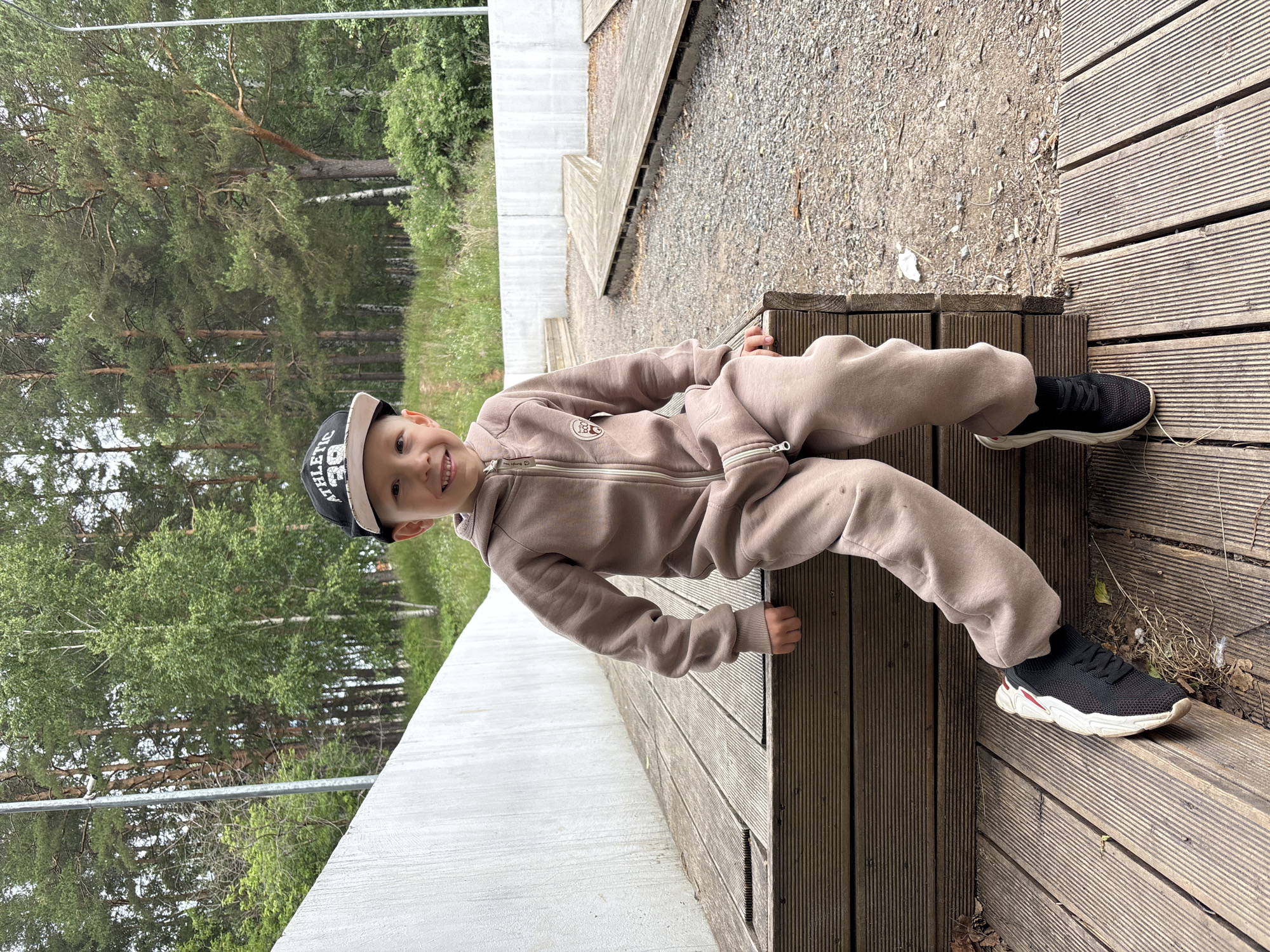 A young boy with fair skin, light brown hair, and blue eyes is sitting on a wooden platform outdoors. He is wearing a light brown hooded jumpsuit with a zipper and a black baseball cap with the word "ATHLETIC" and the number "39" on it. The boy is smiling and looking at the camera. He is wearing black athletic shoes with white soles. The background consists of a concrete wall, trees, and a gravel path with wooden walkways. The scene appears to be in a park or outdoor recreational area. The location is the city of Chaikovsky, Russia.