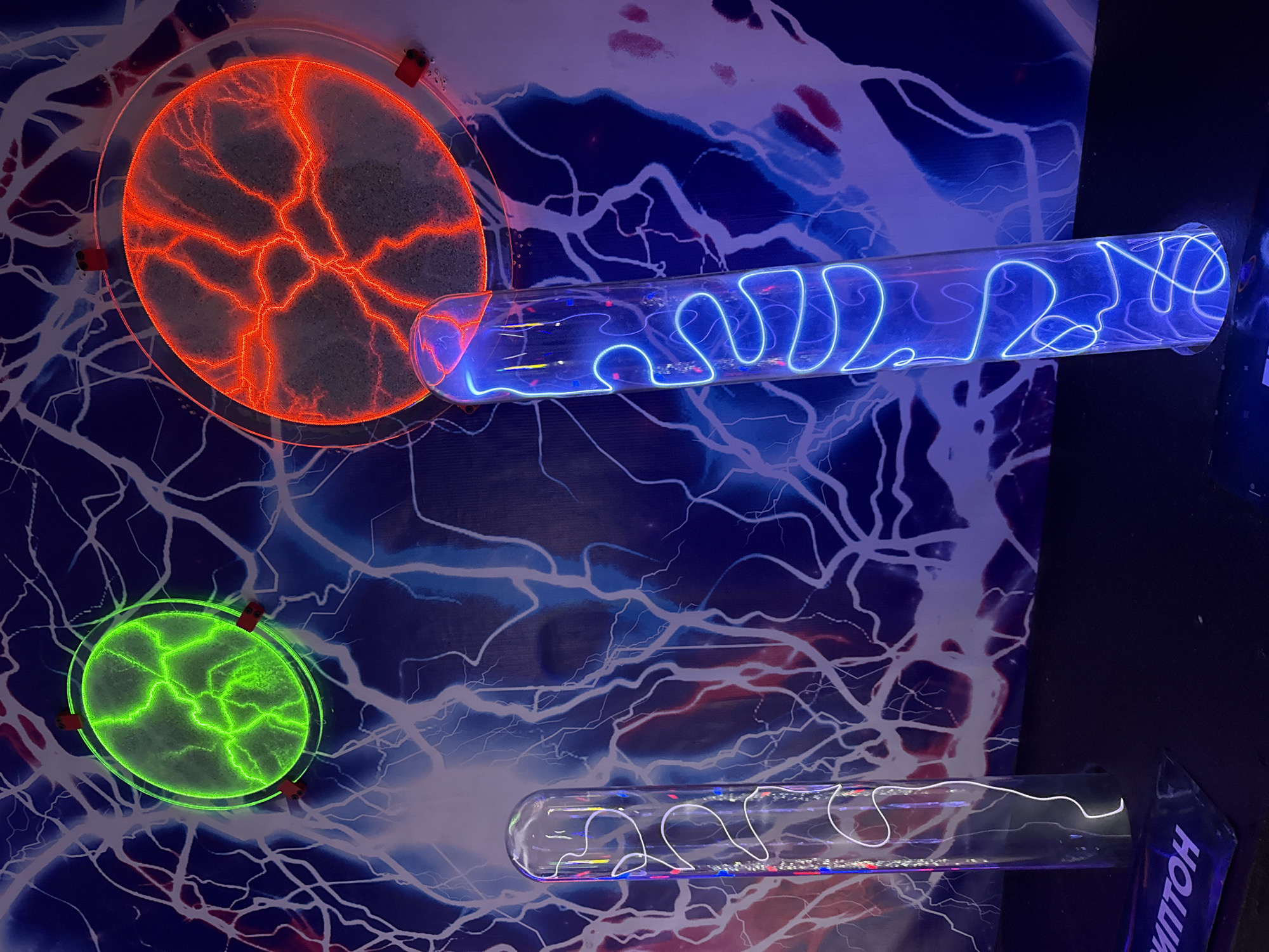 The image shows a display featuring plasma lamps in Kazan, Russia. There are two disc-shaped plasma lamps, one red and one green, and two cylindrical plasma tubes containing blue and white plasma, respectively. These are set against a backdrop with a pattern of branching, lightning-like lines.