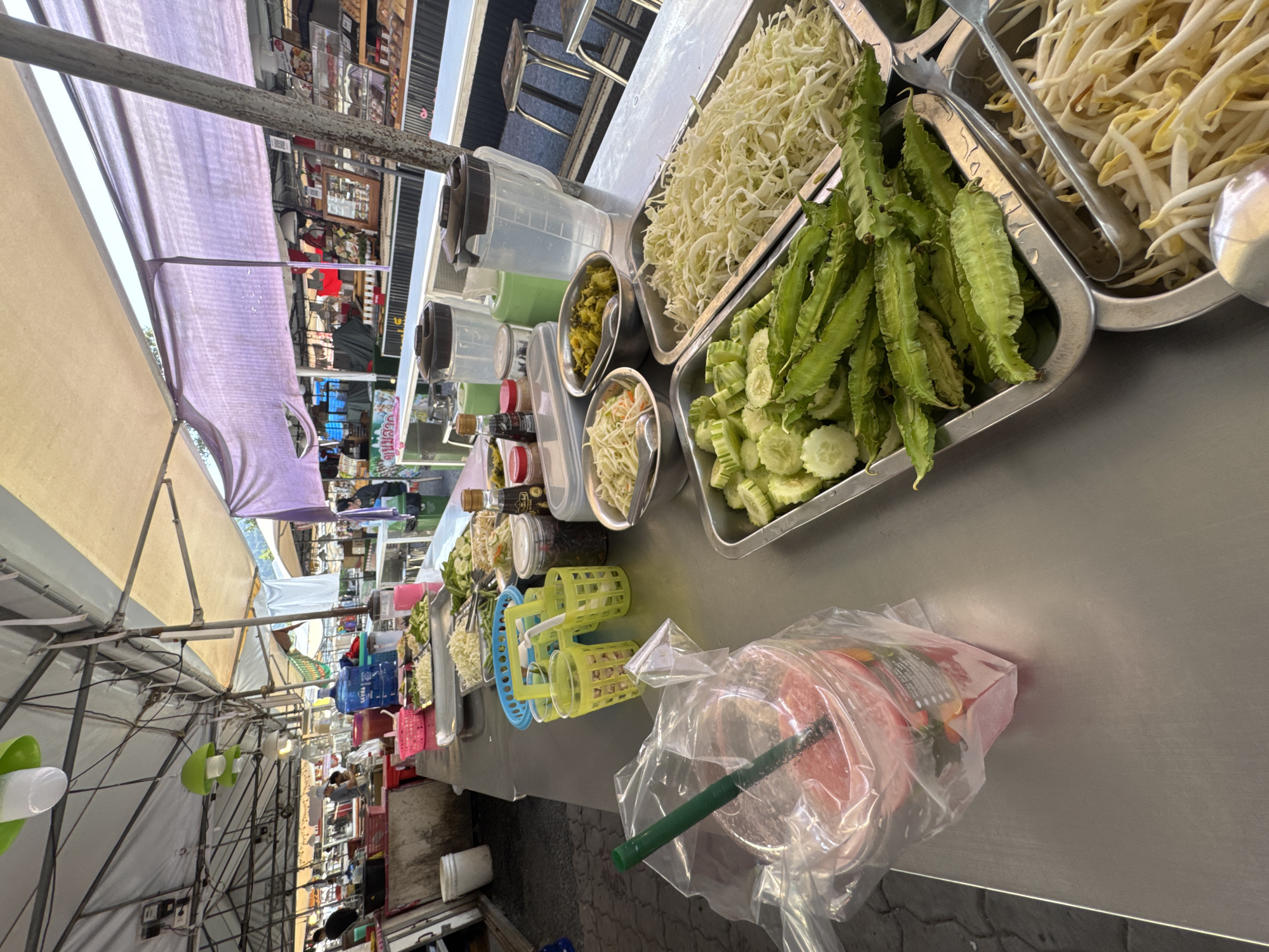 The image shows a food stall in the city of บ้านบางรักษ์, Thailand. The stall's metal countertop displays various ingredients for a dish, including shredded cabbage, bean sprouts, sliced cucumbers, and winged beans in metal trays. There are also smaller bowls with other ingredients and several plastic containers. A plastic cup filled with a red liquid, possibly a drink, and a green straw is in the foreground, encased in a plastic bag. In the background, under the tent-like covering, are other stalls and people, suggesting a bustling market environment. The view is slightly tilted.