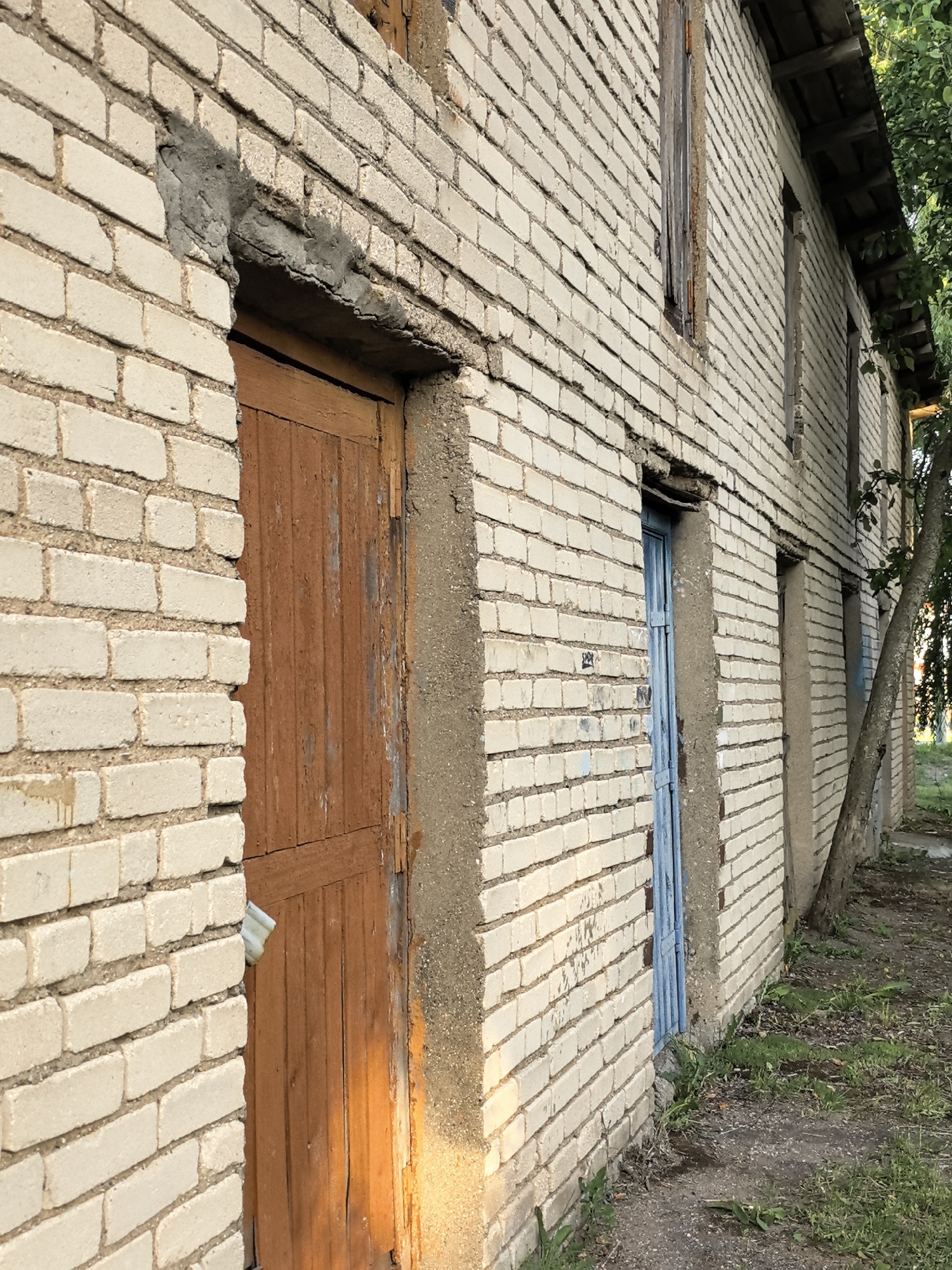 An exterior shot captures the side of a weathered, pale brick building with a sloped roof. Two doors are visible: one on the left is wooden and painted a dull brown, while another further down the wall is painted blue. The building's facade shows signs of age, with chipped mortar and uneven brickwork. A thin tree trunk grows close to the right side of the building, its branches extending upwards. The ground in front of the building is covered with dirt and patches of green grass and weeds. The scene appears to be in a rural or neglected urban setting.