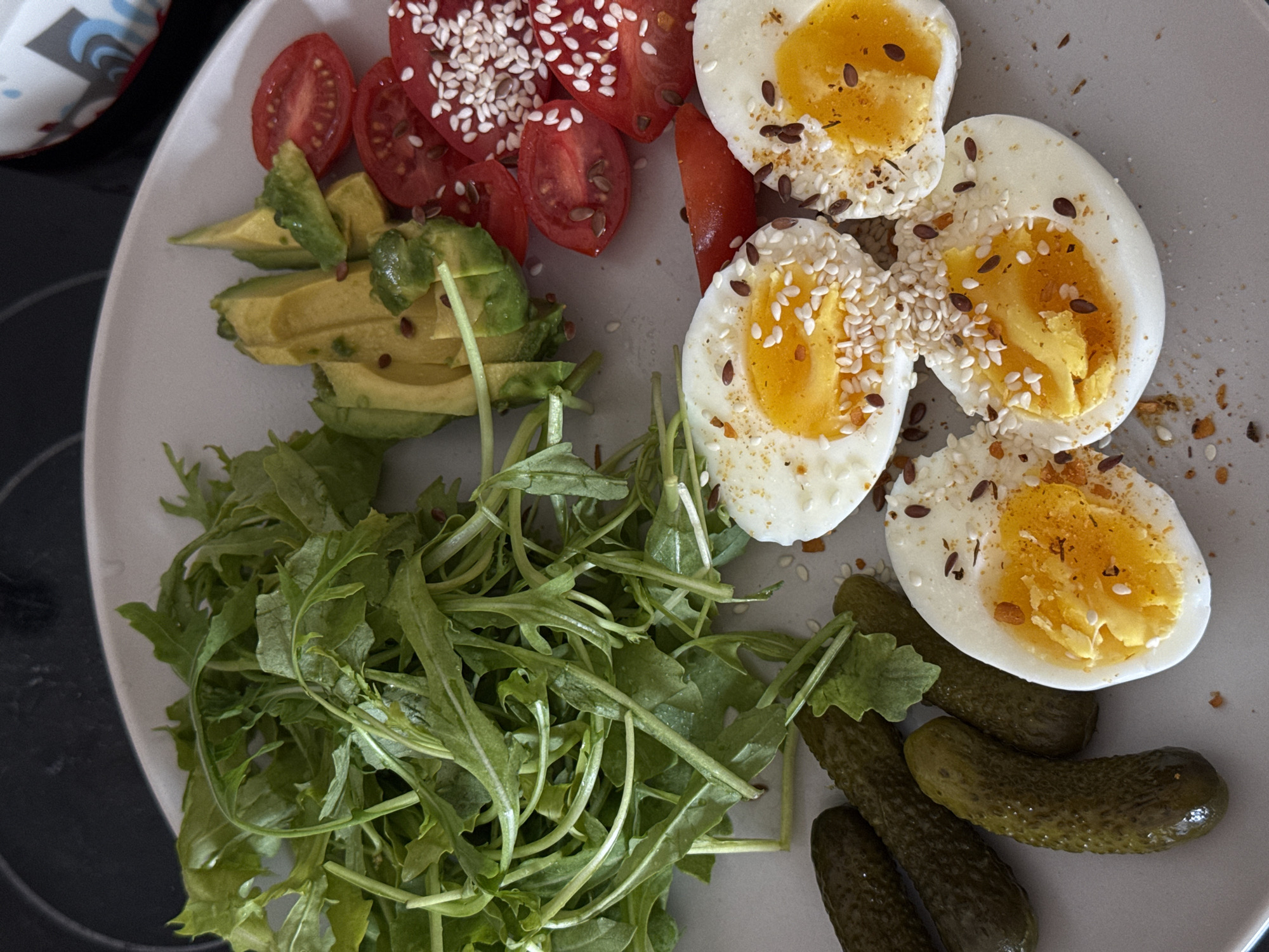 A plate of food is presented. The plate contains arugula salad, sliced avocado, sliced cherry tomatoes with sesame seeds, halved soft-boiled eggs with sesame seeds, and pickles. The scene is likely indoors, with part of a mug visible in the upper left corner. It's impossible to verify the location as Tel-Aviv, Israel from the image alone.